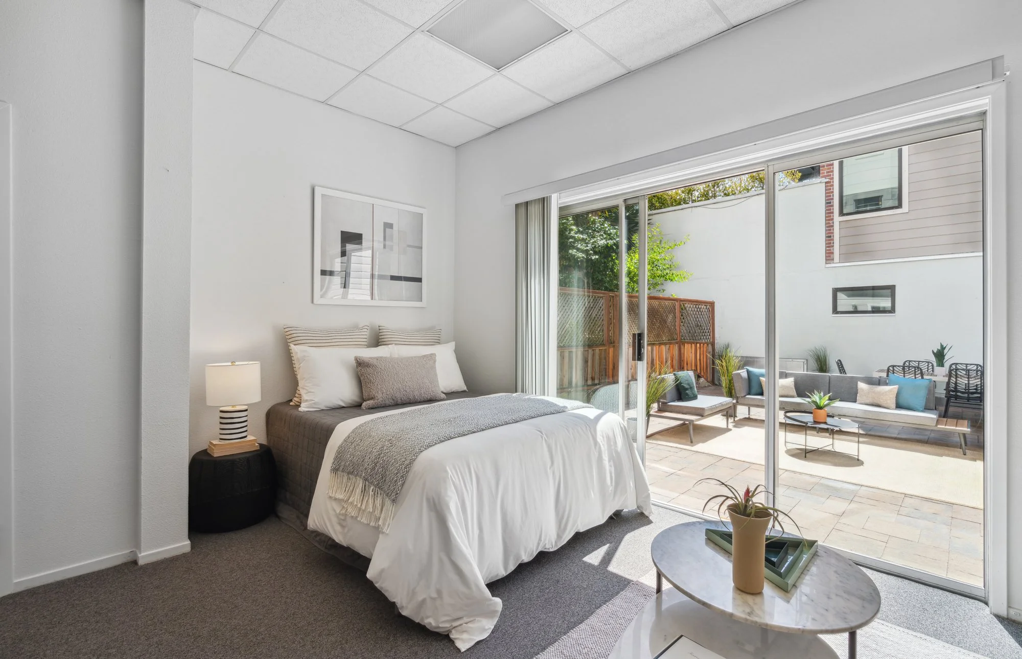 A bedroom with white walls and a bed topped with white and gray bedding, beside a side table with a striped lamp. Large sliding glass doors open to a patio with outdoor furniture and greenery.
