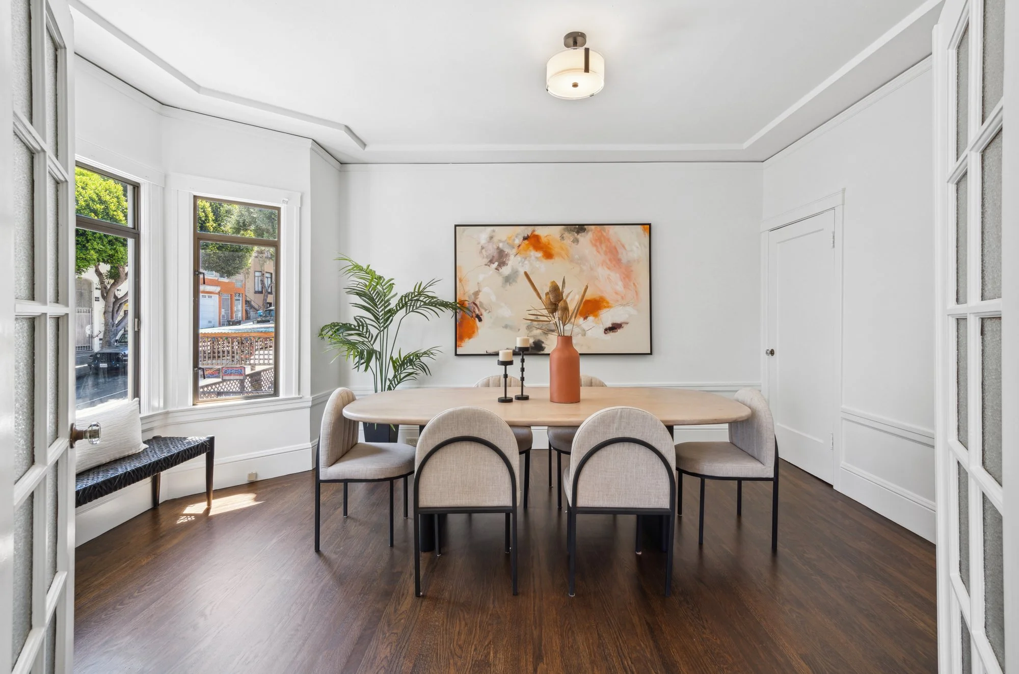 Bright dining room with white walls and large windows, round wooden table with six cream-colored chairs, floating artwork with orange, black, and beige abstract patterns, potted plant, and a terracotta vase with dried plants.
