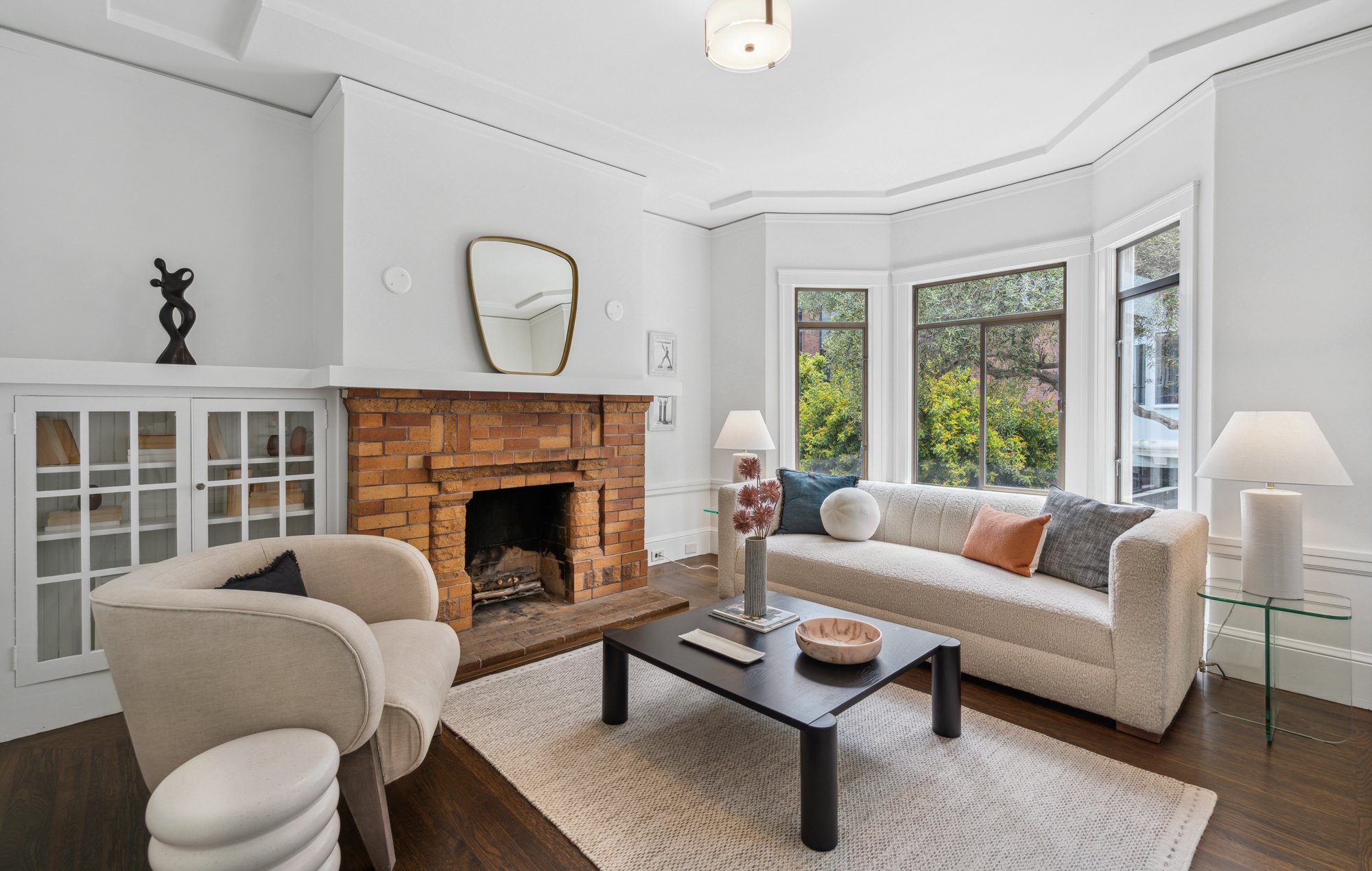 Living room with white walls, a brick fireplace, large windows showing greenery outside, a beige sofa with colorful pillows, a white armchair, black coffee table, and modern art decorations.