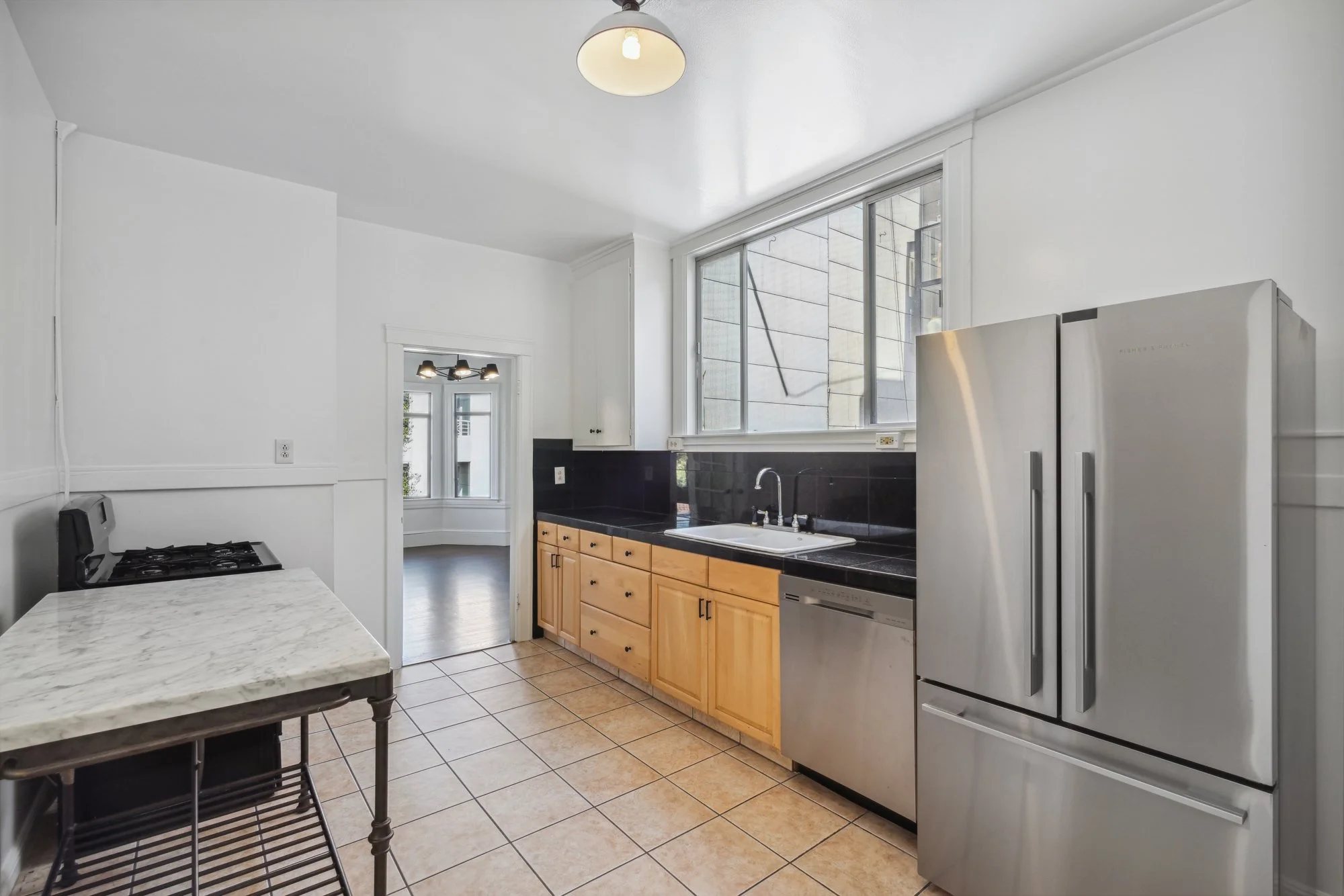 A kitchen with white walls, large windows, wooden cabinets, and stainless steel appliances. There is a small table with a marble top and black chairs in the foreground. The floor is tiled with tan tiles.