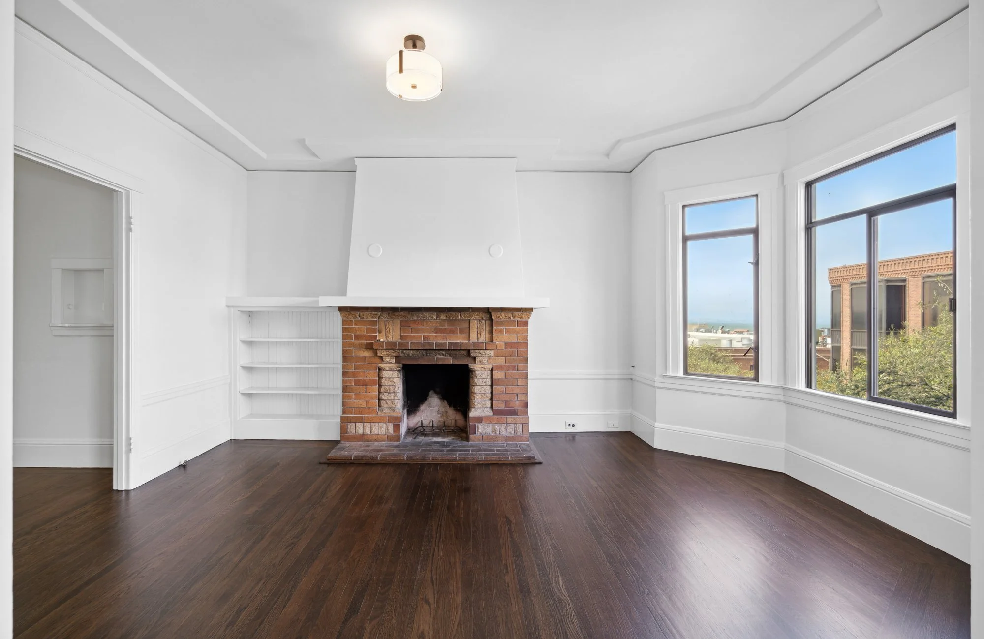Empty living room with white walls, brick fireplace, and large windows showing a cityscape and blue sky.