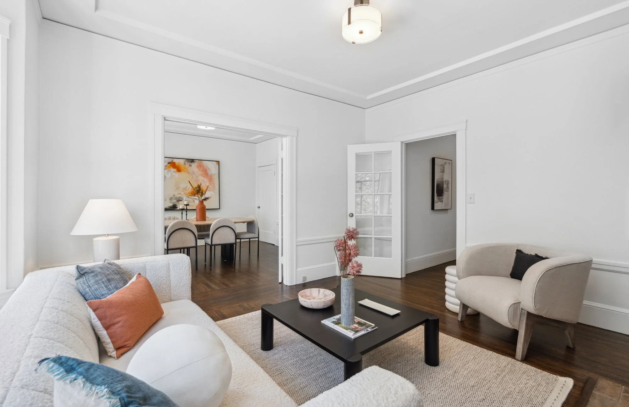 A living room with white walls, hardwood floors, a beige sofa with decorative pillows, a black coffee table with magazines and decor, a beige armchair, a white lamp, and a vase with pink flowers. Visible through an open doorway is a dining area with 