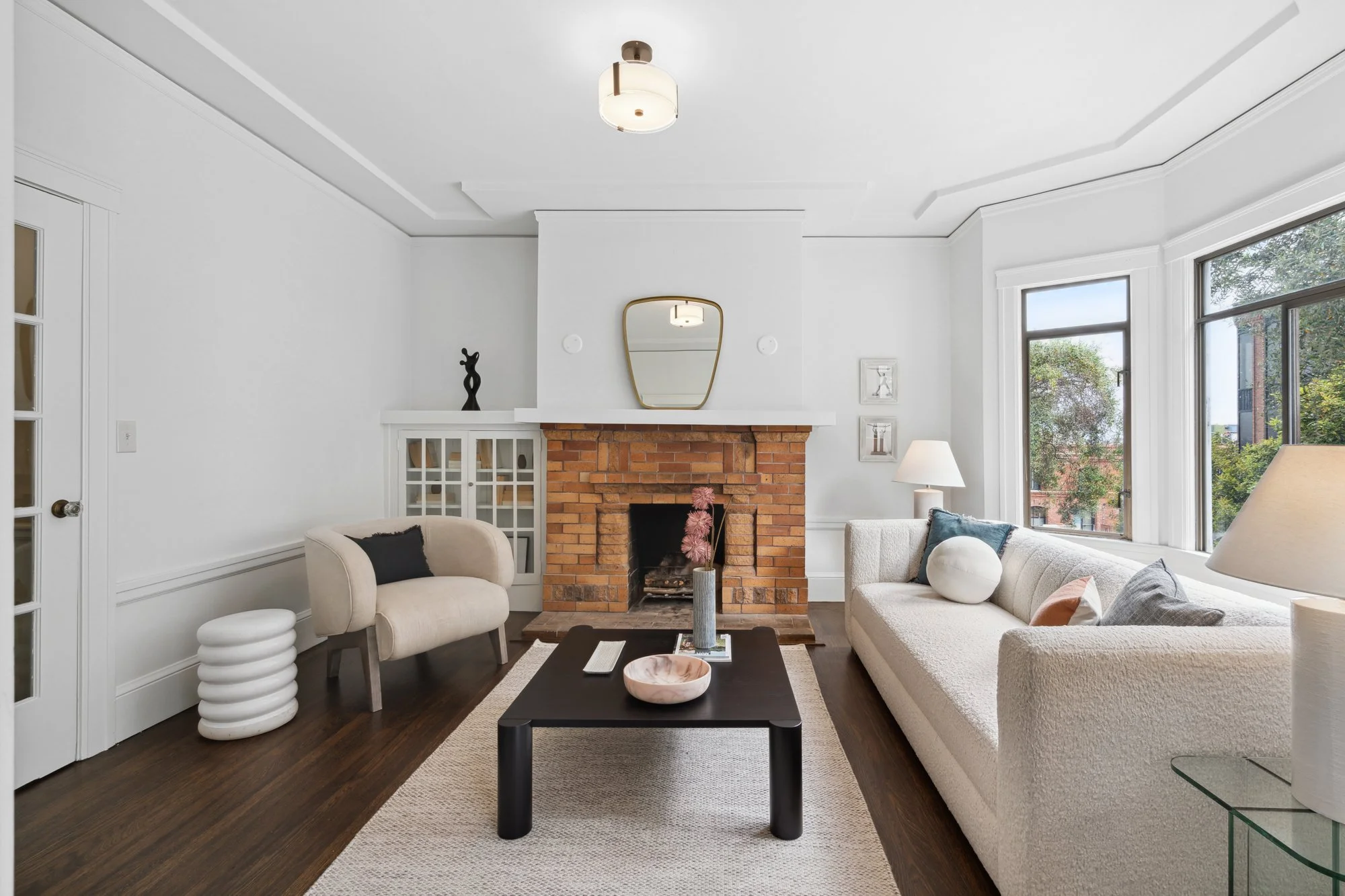 Living room with white walls and large windows, brick fireplace, modern furniture including a white sofa, beige armchair, and black coffee table with decorative bowls, and a white ceiling light fixture.