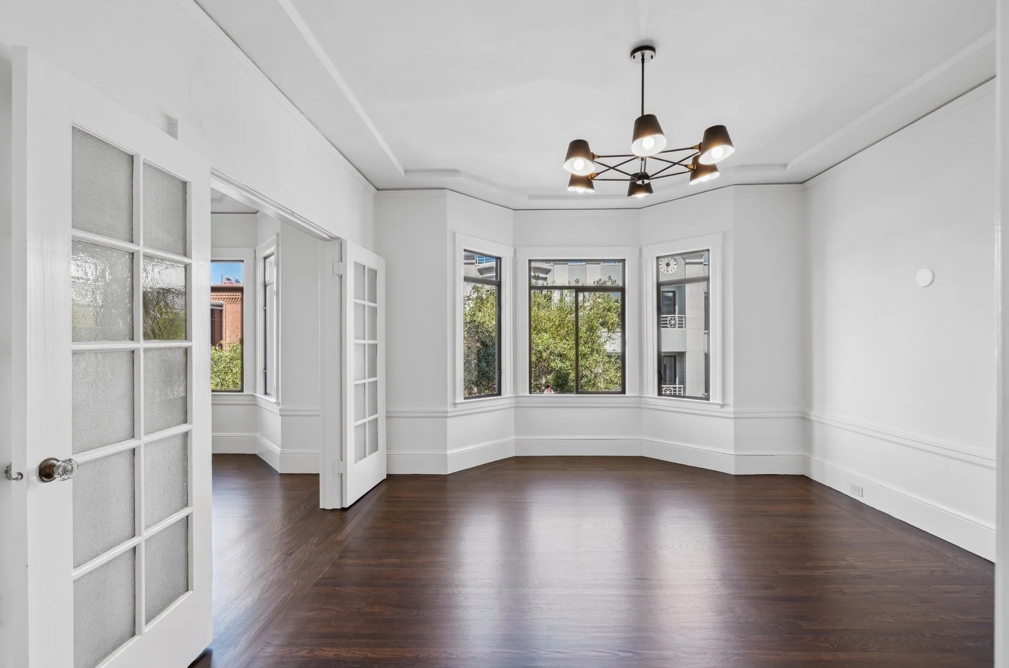 Empty living room with white walls, large bay windows, hardwood floors, and a modern ceiling light fixture.