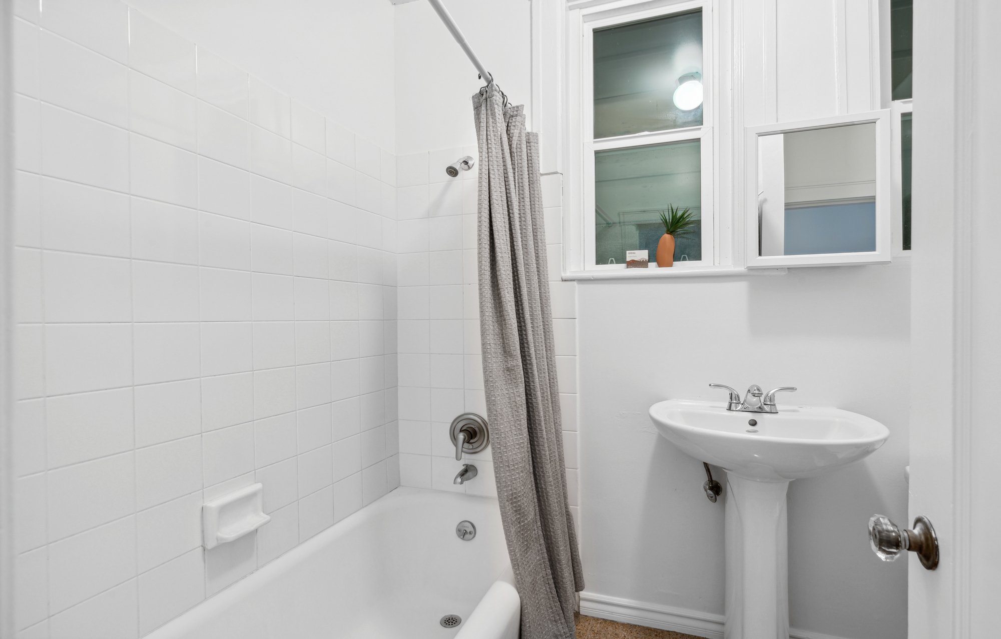 A small bathroom with a white bathtub and shower, a gray shower curtain, a white sink, a mirror, a window with a small potted plant, and a ceiling light.