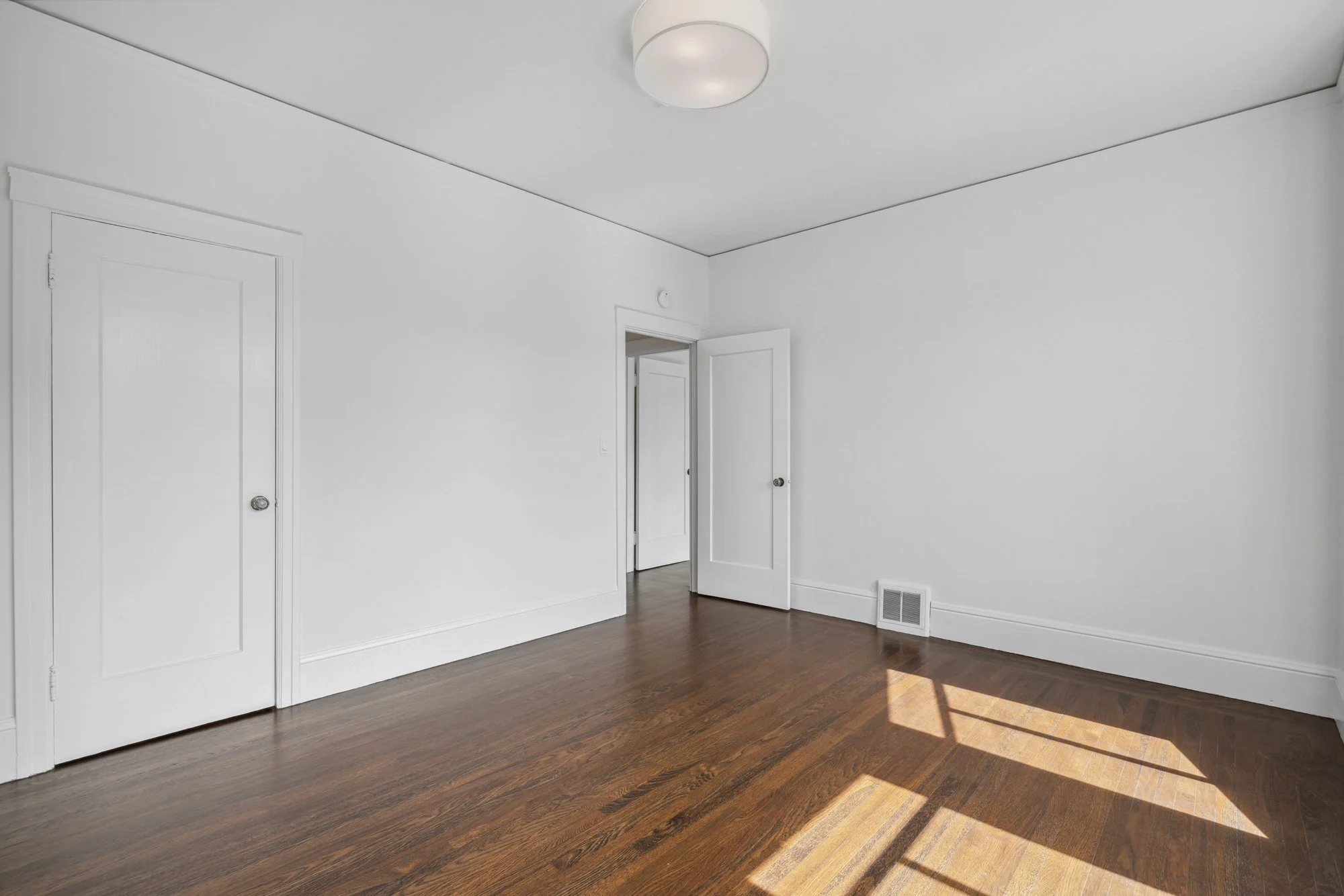 Empty room with white walls, hardwood floor, open door, closed closet door, ceiling light, and a vent near the floor.