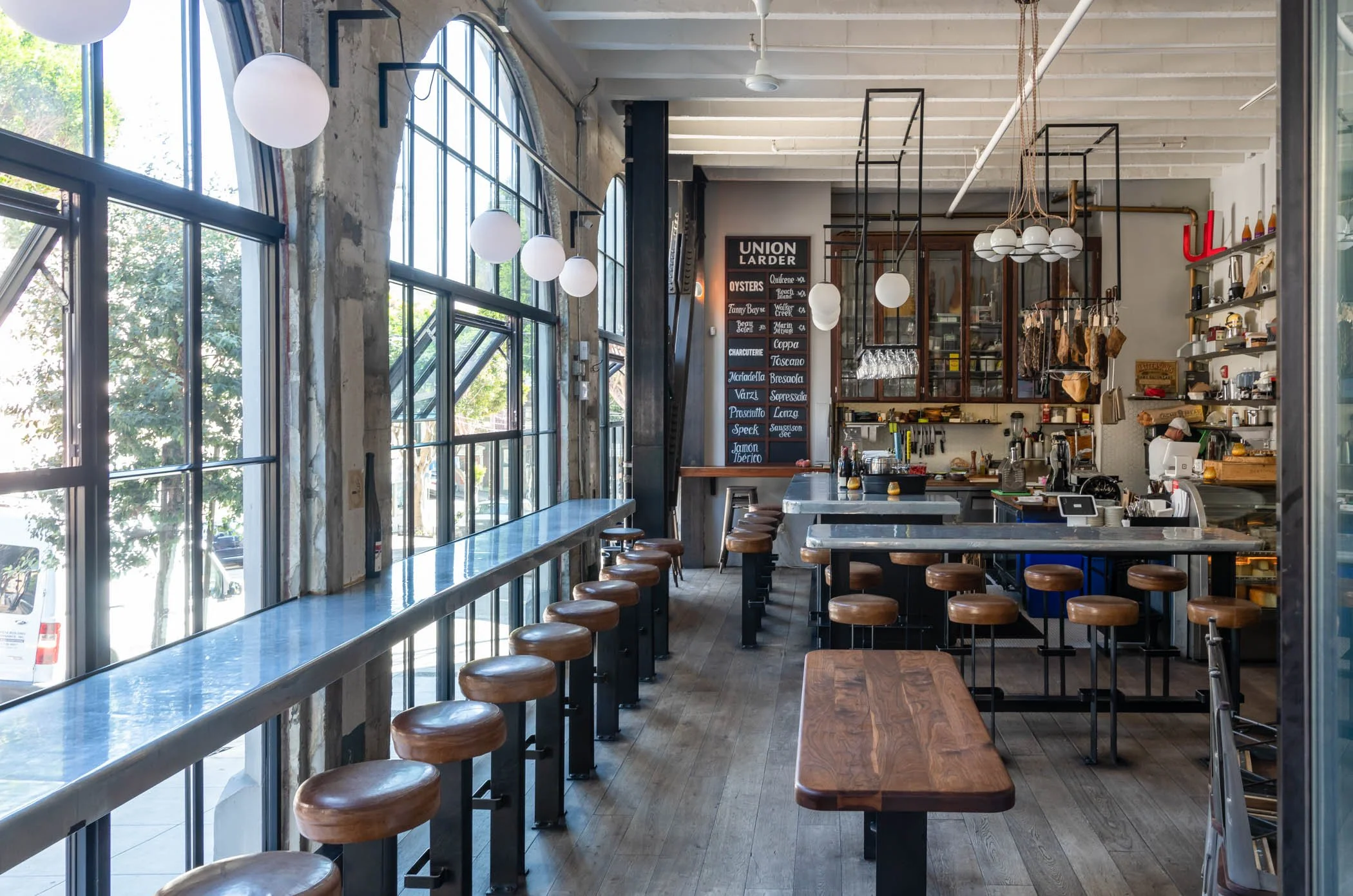 Interior of a modern cafe with large windows, high ceilings, hanging globe lights, a blackboard menu, and minimalist furniture including bar stools and tables.