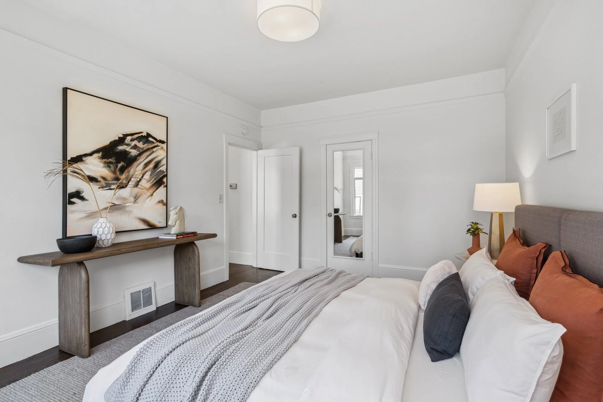 A bedroom with white walls and ceiling, decorated with abstract artwork, a wooden console table with vases, books, and a decorative horse head. A bed with white bedding and rust-colored pillows, a bedside table with a lamp, and a mirror on the wall. 