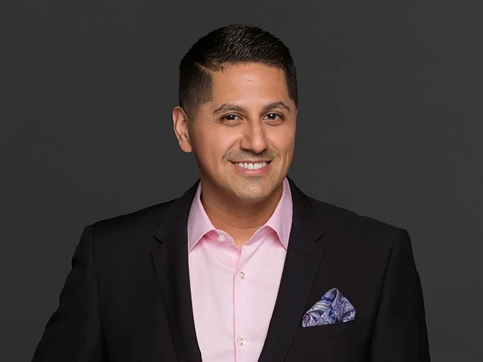 Professional man in a black suit and pink shirt smiling against a dark gray background.