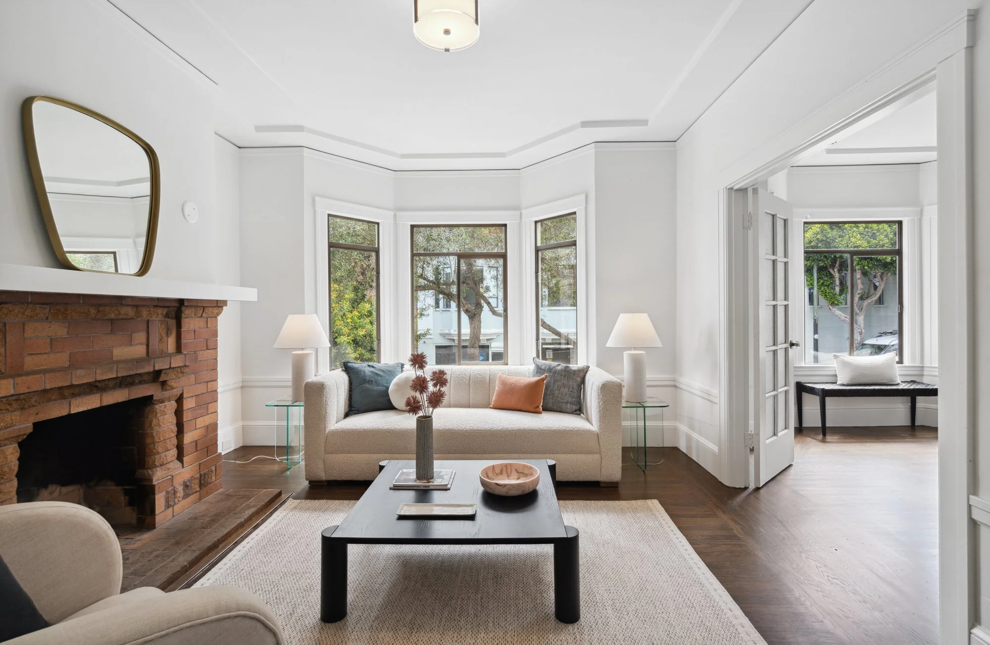 Bright living room with large windows, white walls, a brick fireplace, cream sofa with throw pillows, glass side tables, a black coffee table with decor, and a door leading outside, with trees visible through the windows.