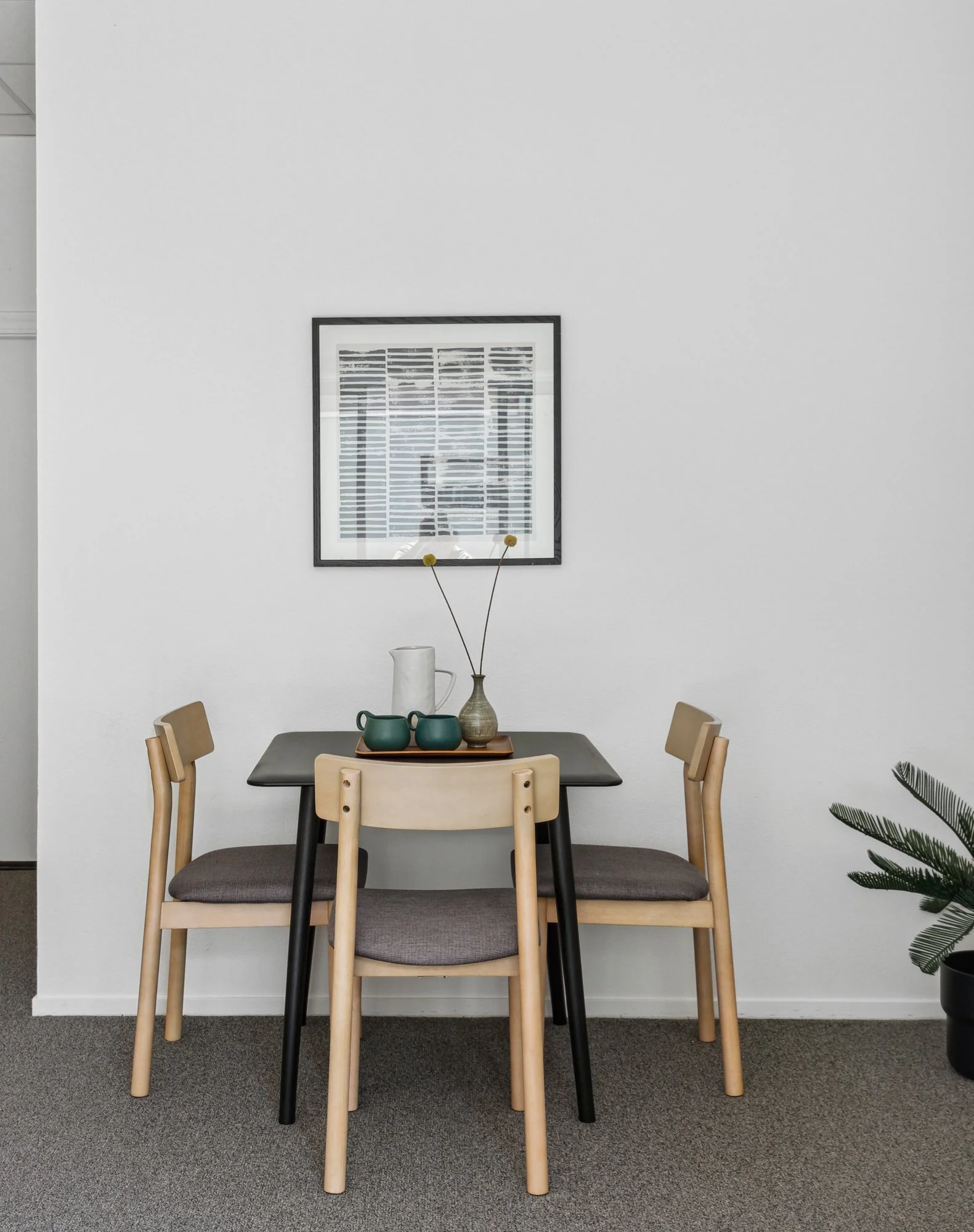 A modern dining room with a black table, surrounded by four wooden chairs with gray cushions. On the table are three dark green cups, a white pitcher, and a beige vase with two yellow flowers. The white wall behind has a framed black-and-white abstra