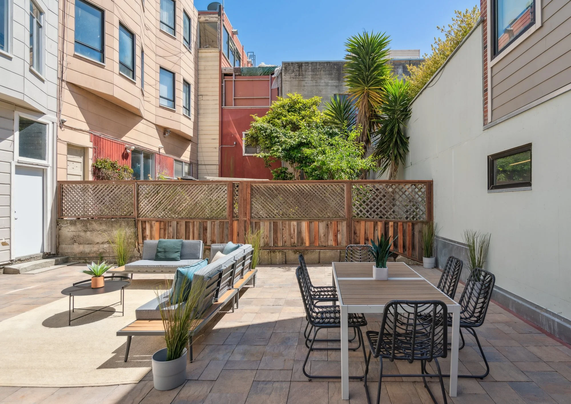 A backyard patio with outdoor furniture, potted plants, and a fenced border, surrounded by multi-story residential buildings and trees under a clear blue sky.