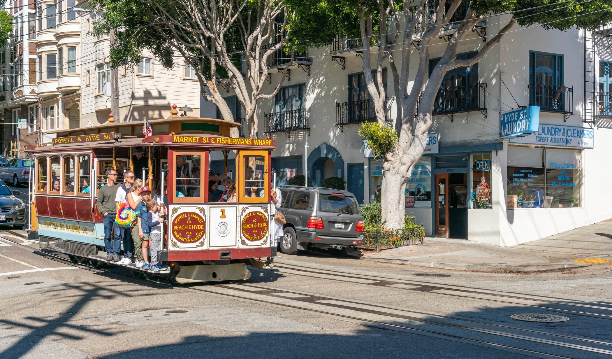 A San Francisco cable car with passengers riding, heading past storefronts including a laundry and barber shop, on a sunny street lined with large trees.