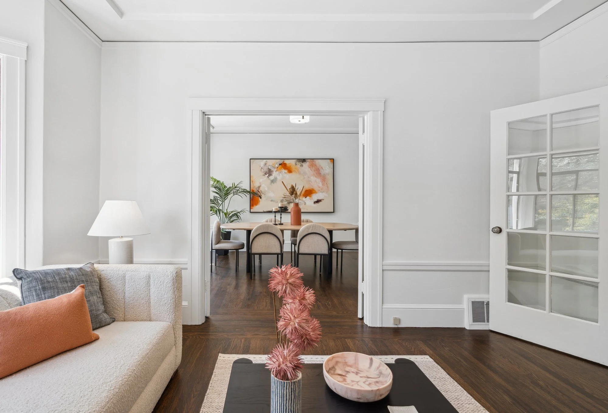A living room with a beige couch with pillows, a white lamp, a coffee table with pink flowers in a striped vase and a marble bowl, and a view into a dining area with a wooden table, beige chairs, a large abstract painting, and a green plant.