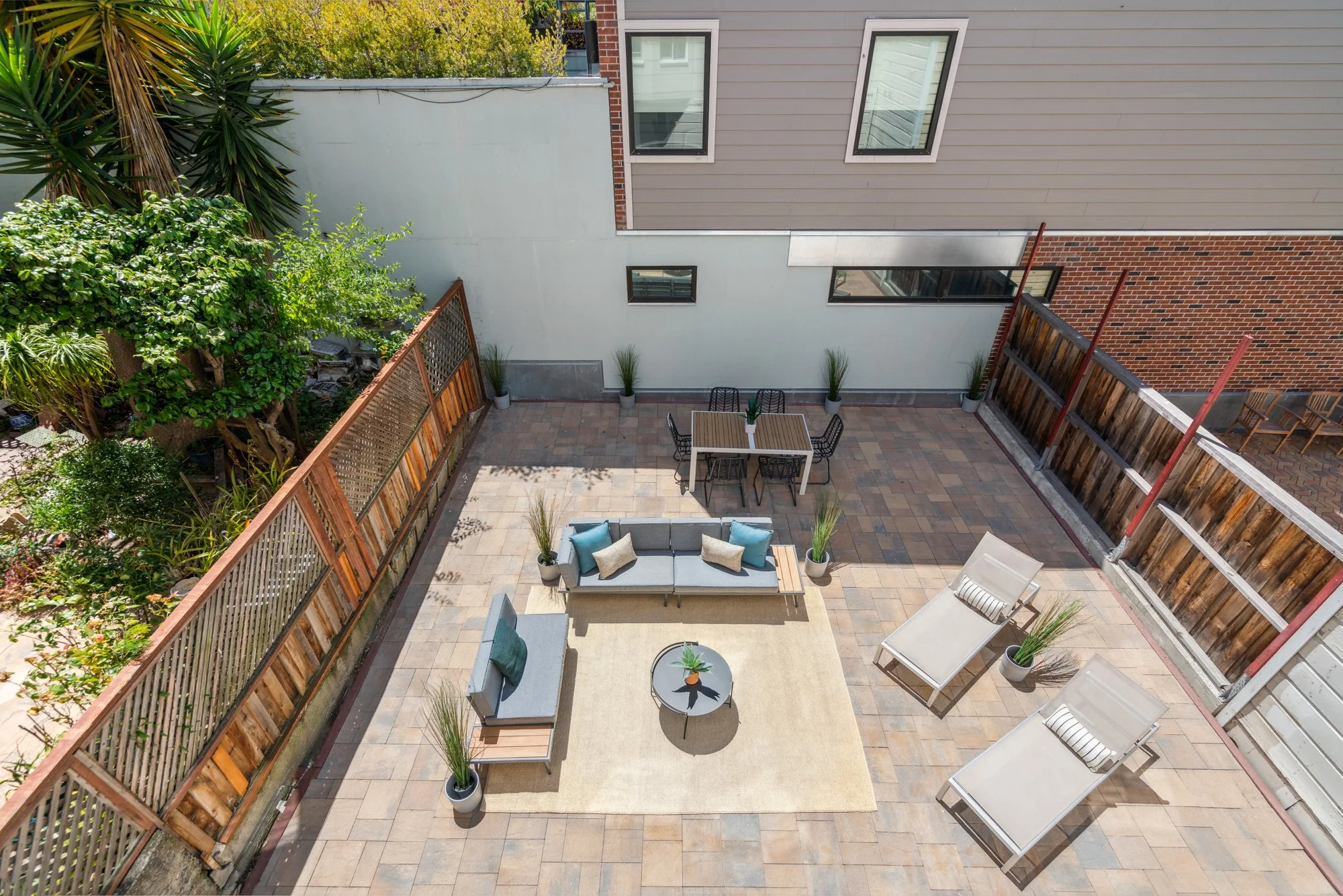 A spacious backyard patio with outdoor furniture, including a sofa, chairs, and a round table with a small potted plant. The patio is enclosed by wooden fences, adorned with potted plants, and has a brick and gray siding house with multiple windows v