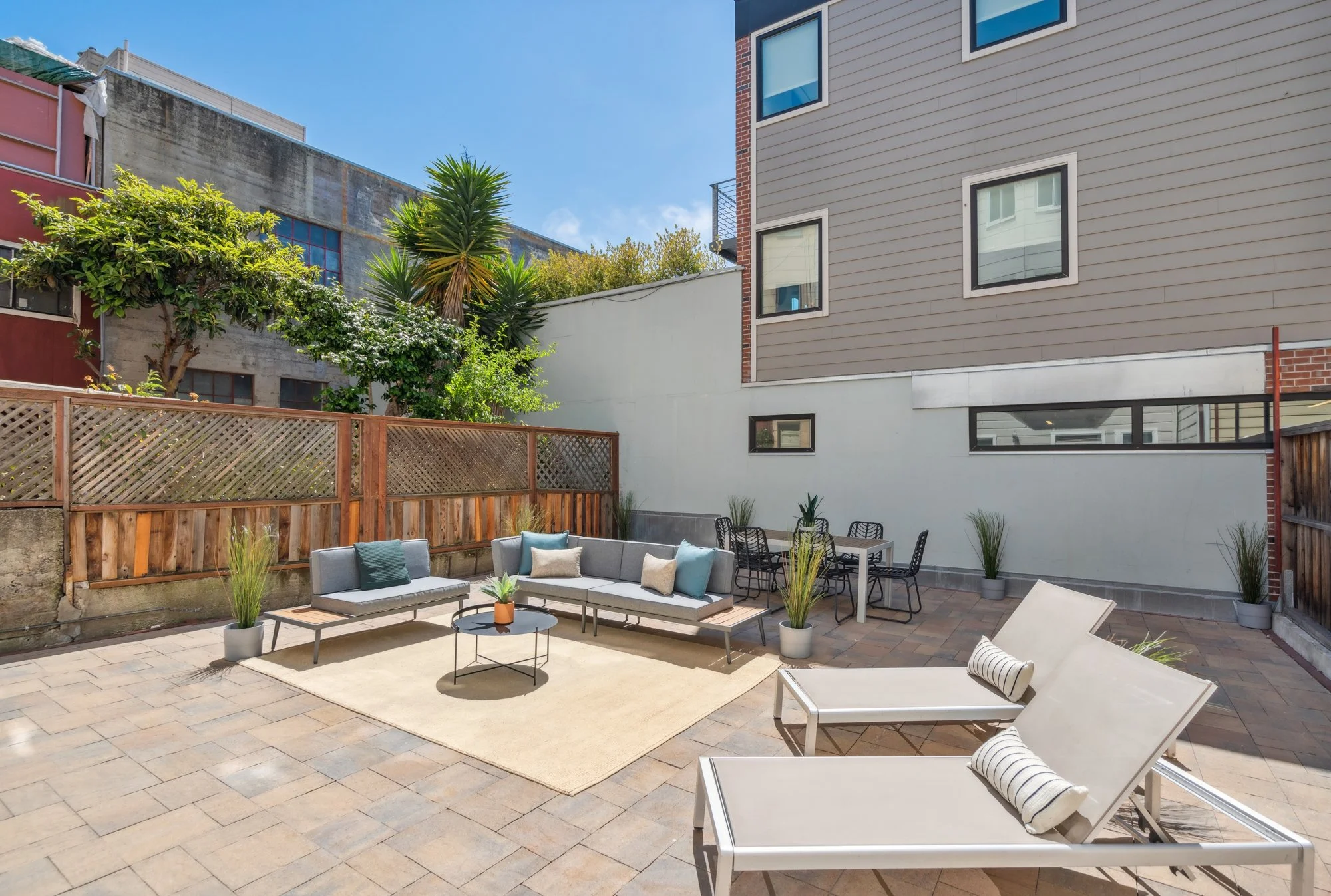 A backyard patio with outdoor seating and lounging furniture, including two white lounge chairs with striped cushions, a gray sectional sofa with pillows, and a black dining table with chairs. Potted plants are placed around the area on a brick-paved surface, enclosed by a wooden fence. In the background, there is a modern multi-story house with beige siding and several windows, and a white wall with small plants at the base. Tall green trees and neighboring buildings are visible under a clear blue sky.