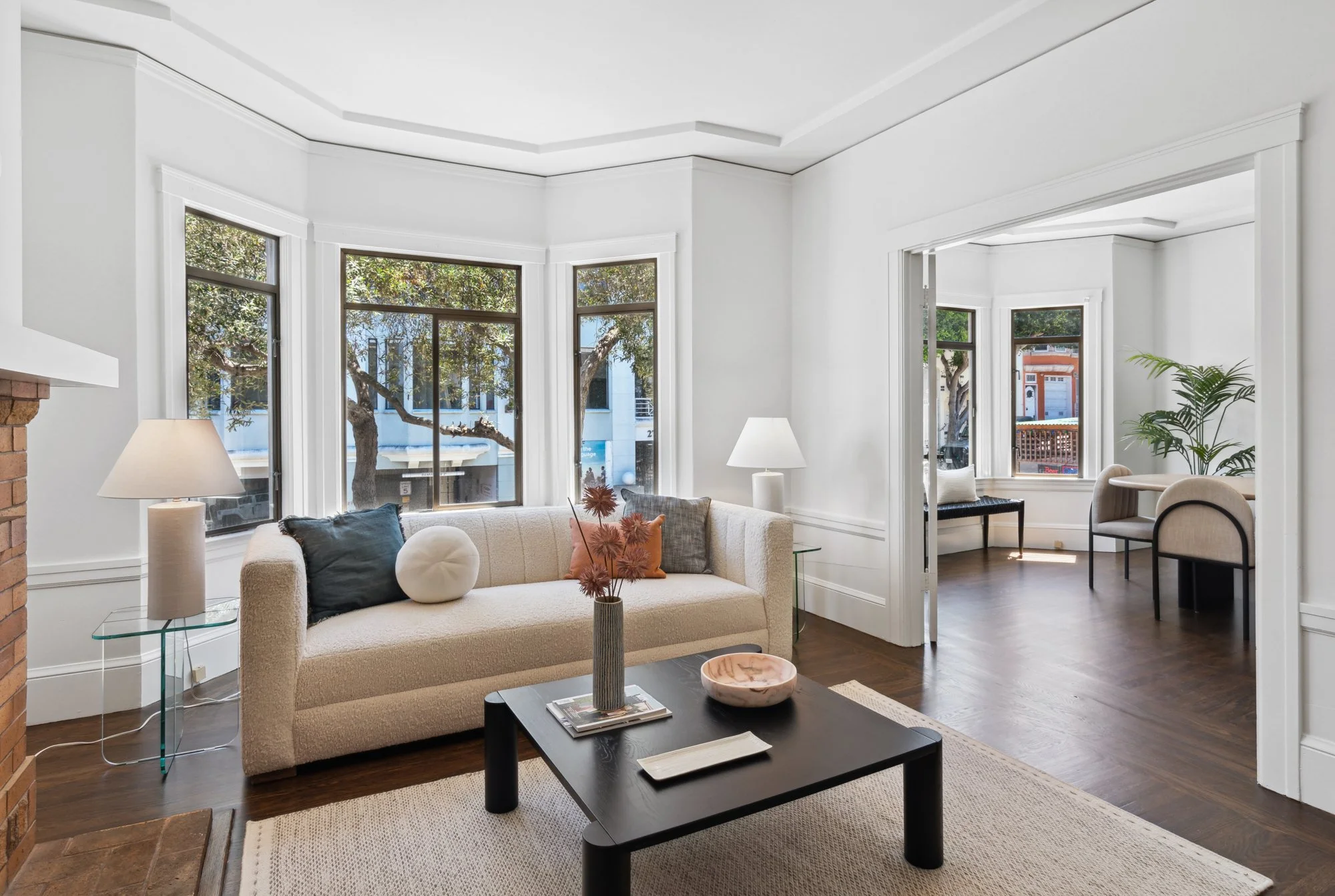 A living room with large windows letting in natural light, decorated with a beige sofa with pillows, a glass side table with a lamp, a coffee table with magazines and decorative items,, a plant, and a doorway leading to a dining area with additional 