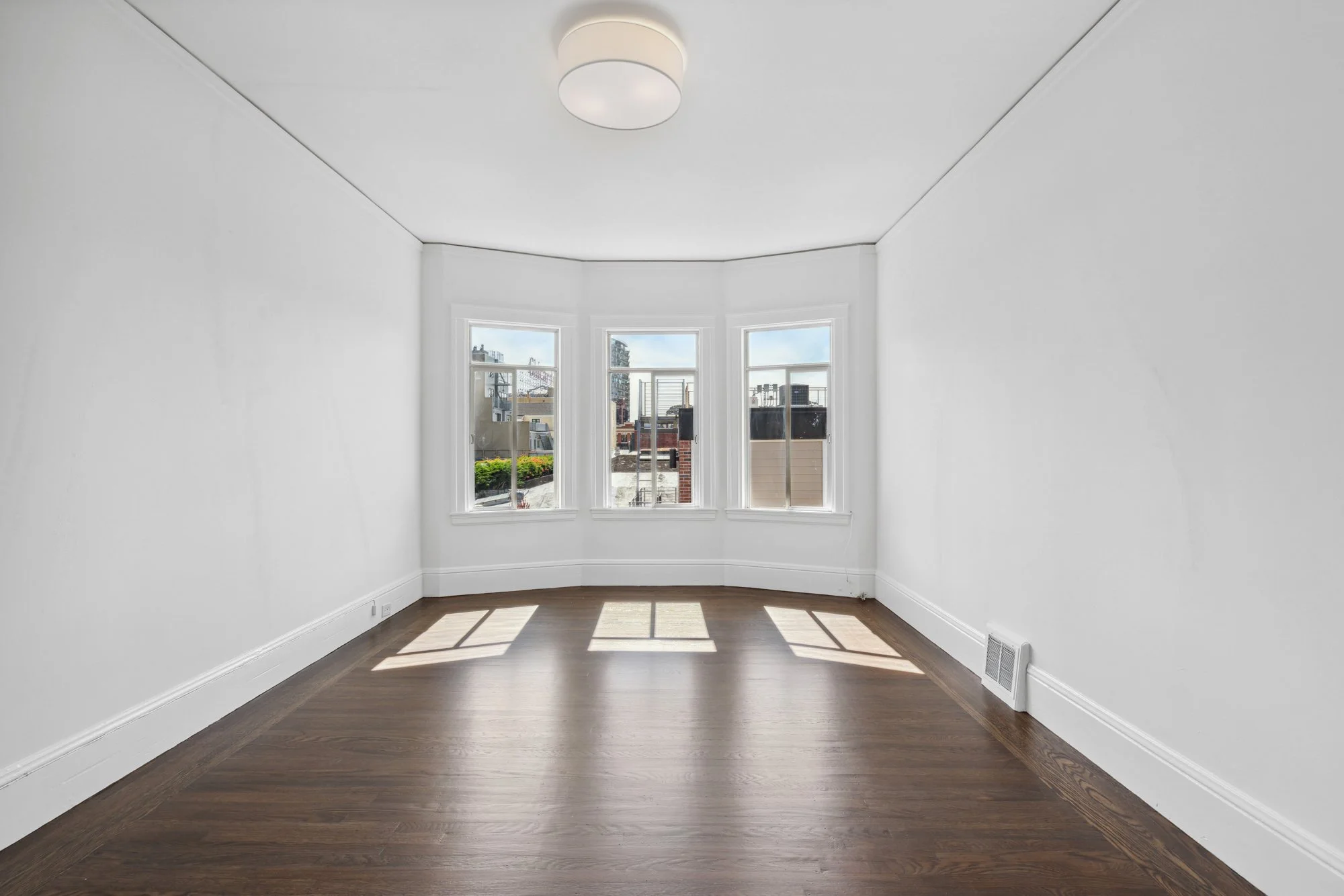 Empty room with white walls, large bay window, hardwood floor, and ceiling light fixture.