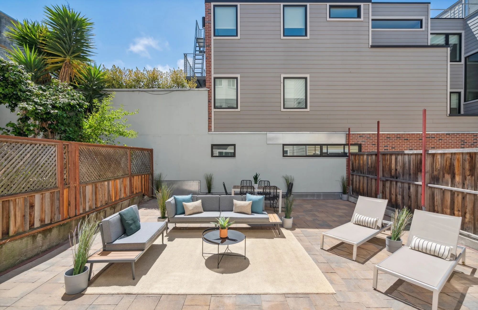 Outdoor patio area with modern furniture, including a sectional sofa with pillows, chairs, a small round table, potted plants, and a beige rug, enclosed by a wooden fence with an urban building in the background.