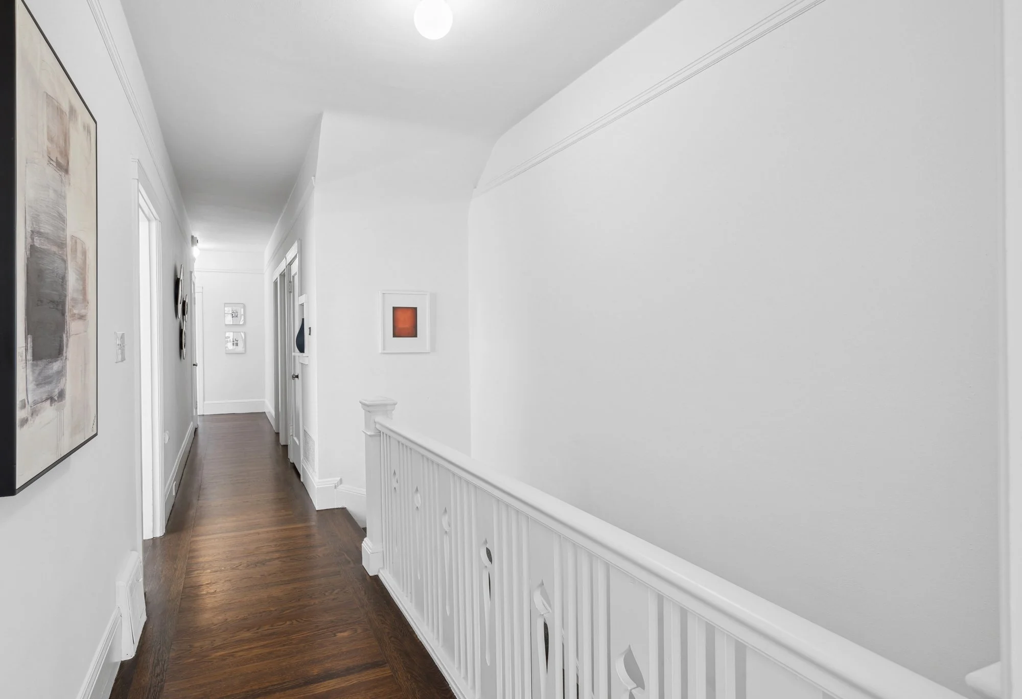 Clean white hallway with wooden floors, decorated with framed abstract artwork on the left wall and smaller framed artworks on the far wall. The ceiling has a single round light fixture, and there is a white wainscoting along the right wall.