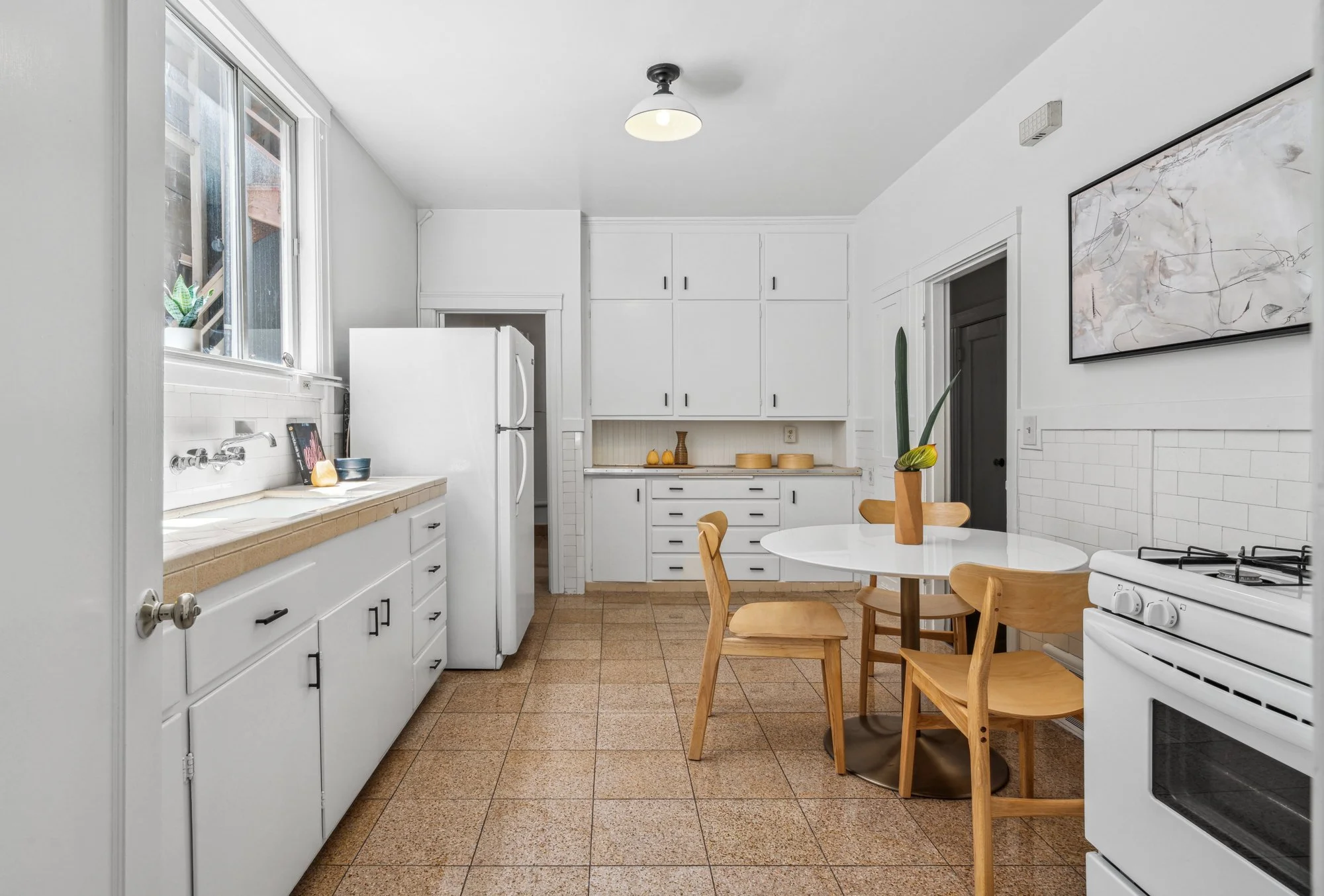 A bright, white kitchen with light wood chairs around a small round table. Cabinets above and below, a window with a small plant on the sill, and a white refrigerator. A large abstract art piece hangs on the wall.
