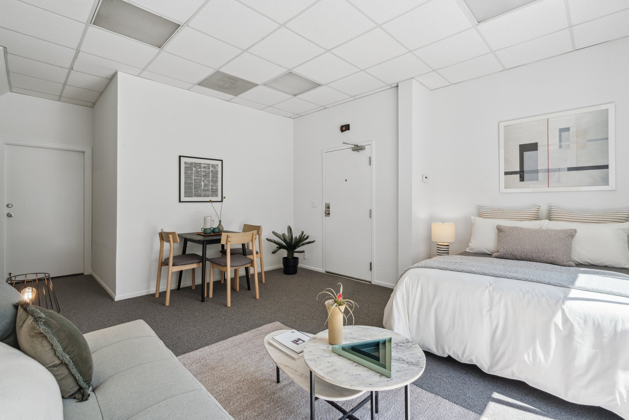 A modern, minimalistic studio apartment with a partial view of a bed, a small dining area with four chairs, a gray sofa, a white marble coffee table with a decorative tray and a plant, and framed artwork on the white walls. The room has a suspended c
