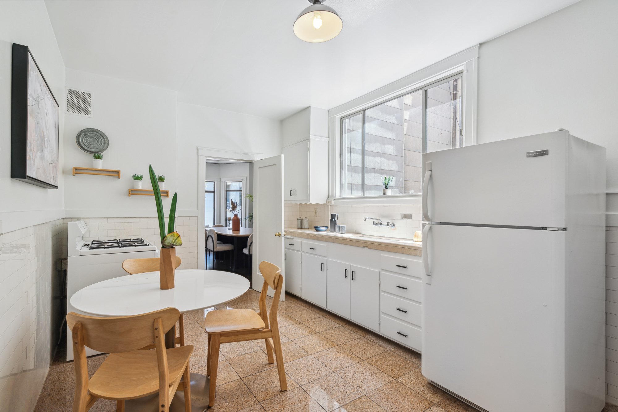 A kitchen with white cabinets, a refrigerator, large windows, a round wooden table surrounded by wooden chairs, and a small dining area visible through an open door.