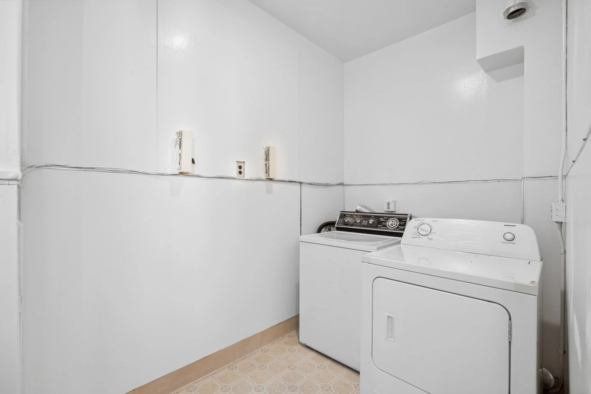 Laundry room with a washing machine and a dryer, white walls, electrical outlets and visible wiring, small window or vent in upper corner.