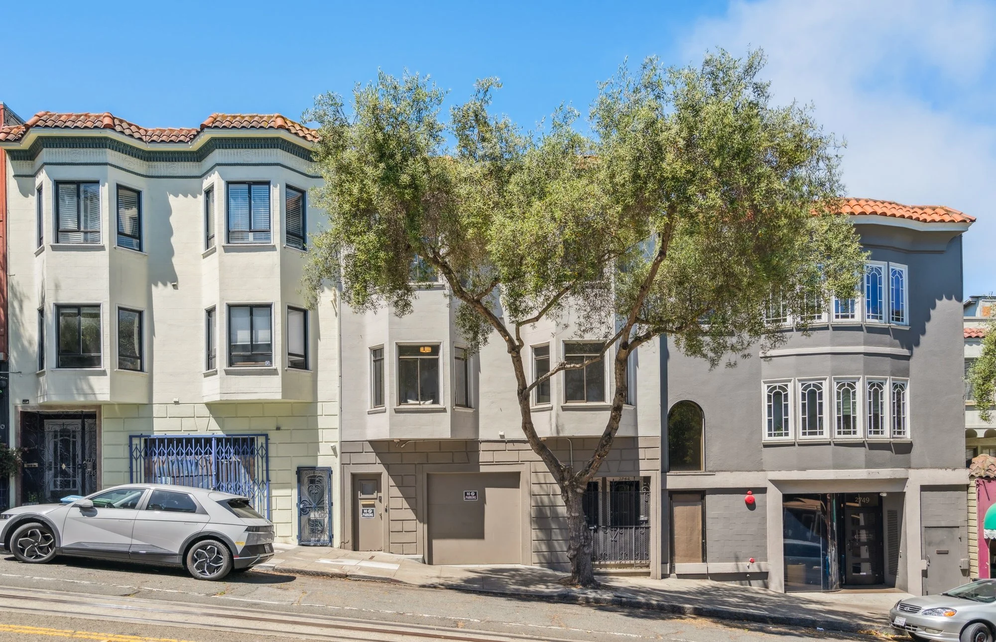 A white residential building with several windows, a tree in front, parked cars, and a street with tram tracks.