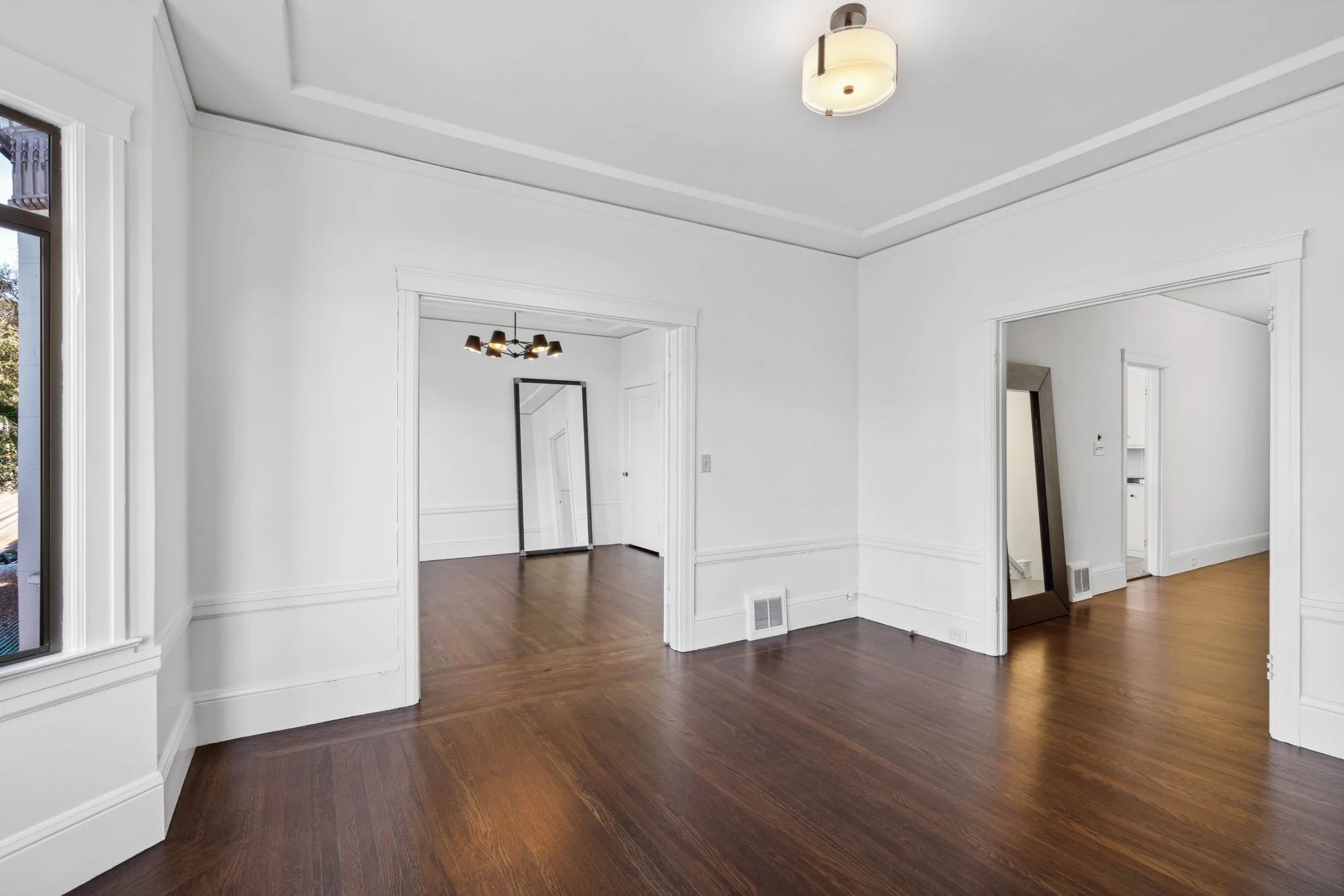 Living room with hardwood floors, white walls, multiple doorways, a large mirror leaning against the wall, and a ceiling light fixture.