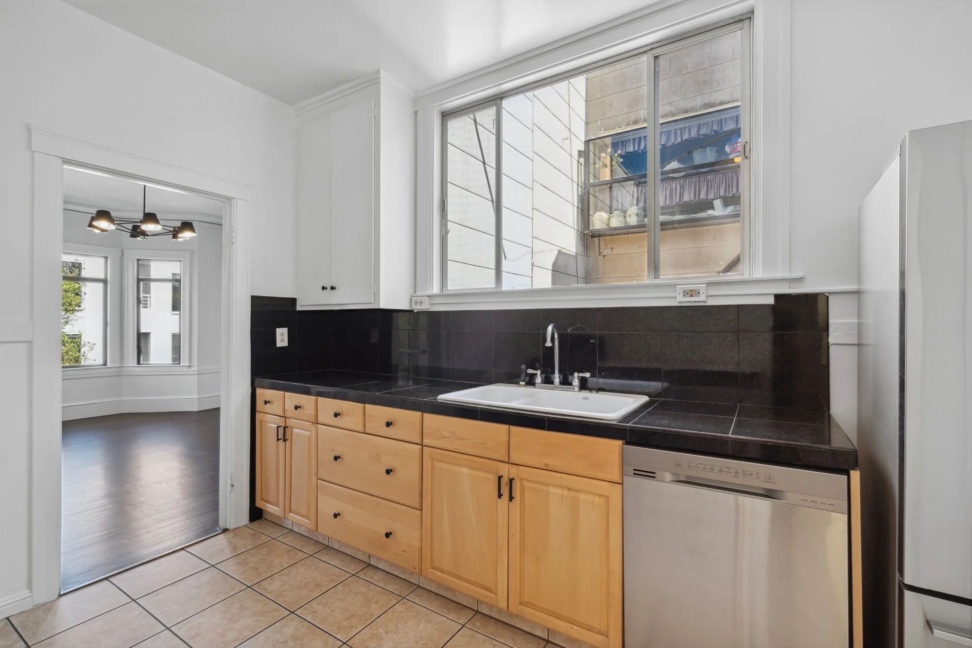 A kitchen with wooden cabinets, black countertop, and white sink under a large window showing outside building and storage with a blue awning.