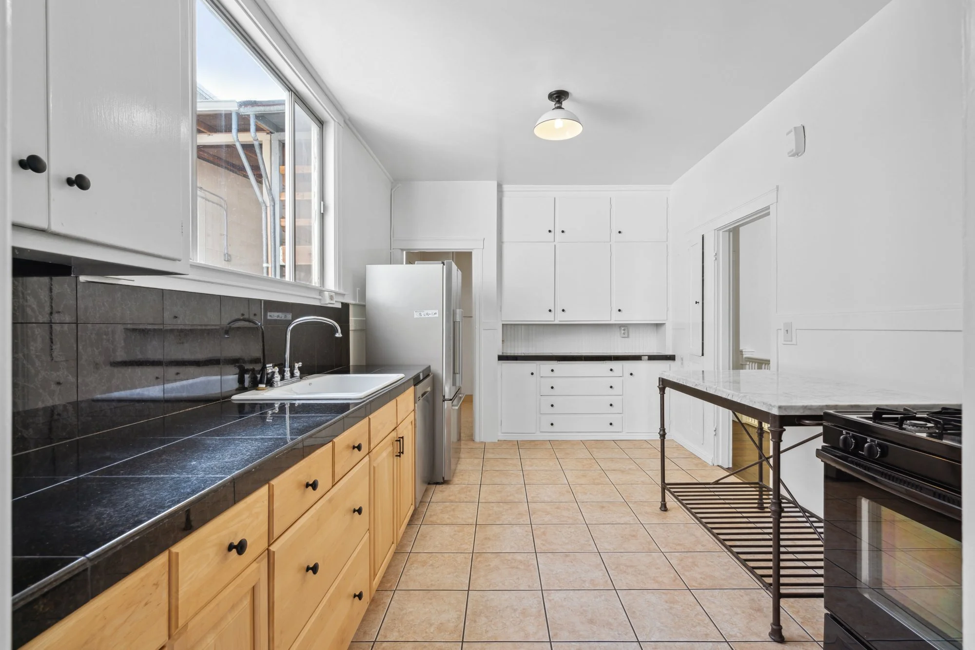 Kitchen with white walls, black tiled countertop, wooden lower cabinets, white upper cabinets, a stainless steel refrigerator, and a black stove with a metal rack nearby. There is a large window letting in natural light.