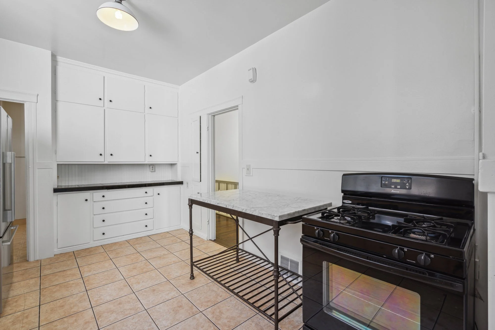A kitchen with white cabinets, a black stove, a marble-top table, and beige tile flooring.