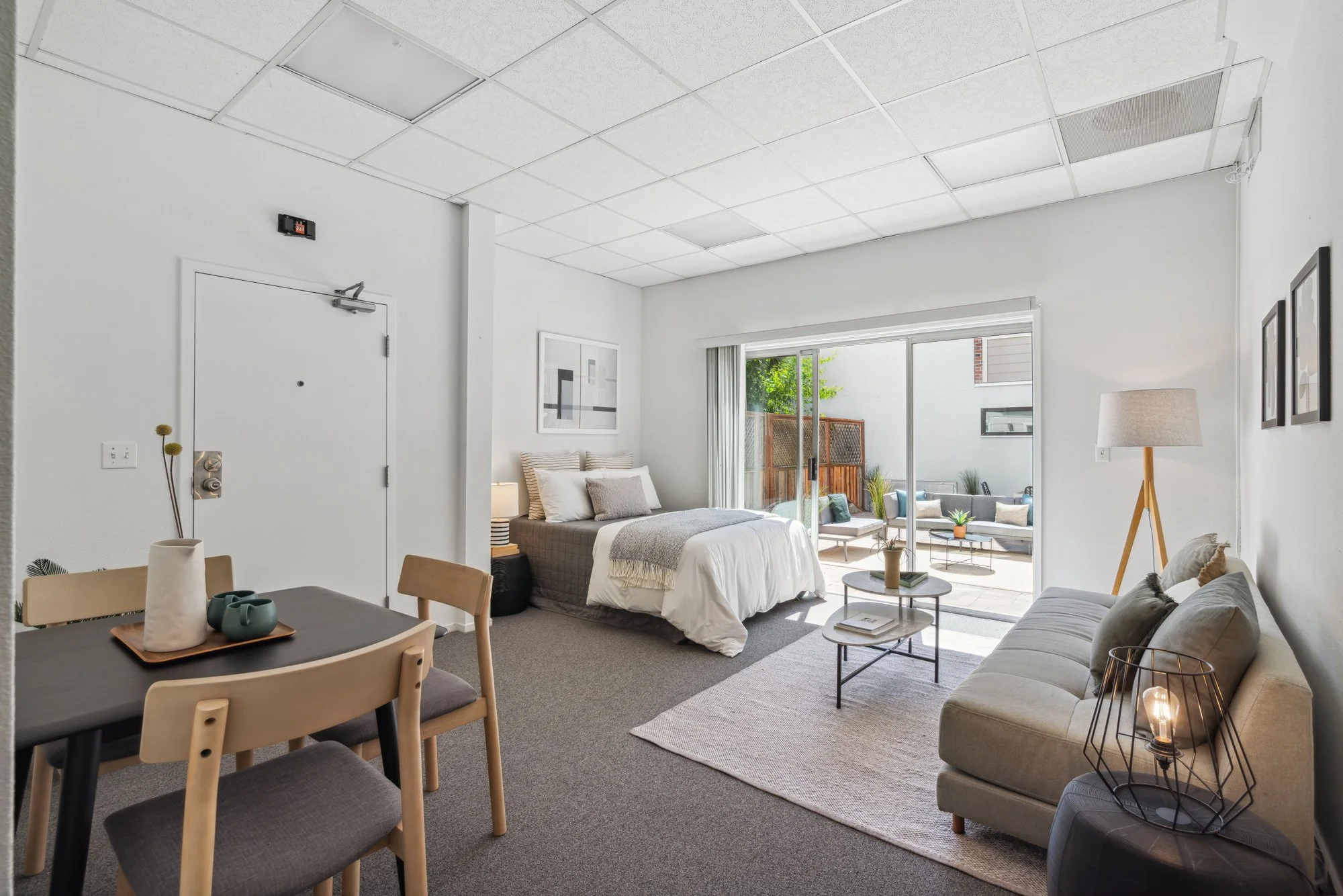 Living room with white walls, a bed near large glass sliding doors, a beige sofa, a round coffee table, a floor lamp, and framed artwork, patio with outdoor seating visible outside.
