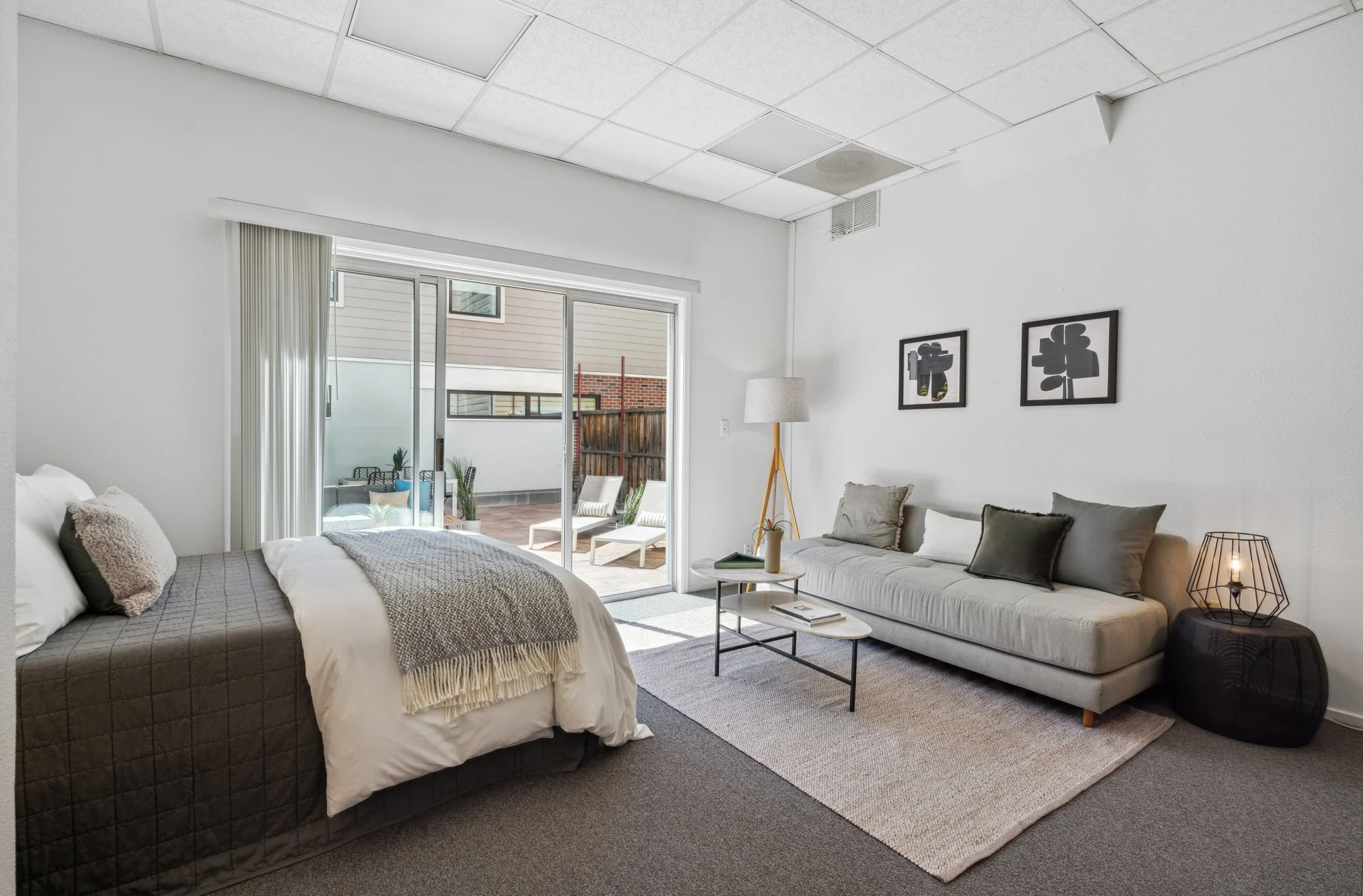 Living room with a bed near sliding glass doors, a gray couch, a white round coffee table, framed art on the wall, and a patio with outdoor chairs visible through the doors.