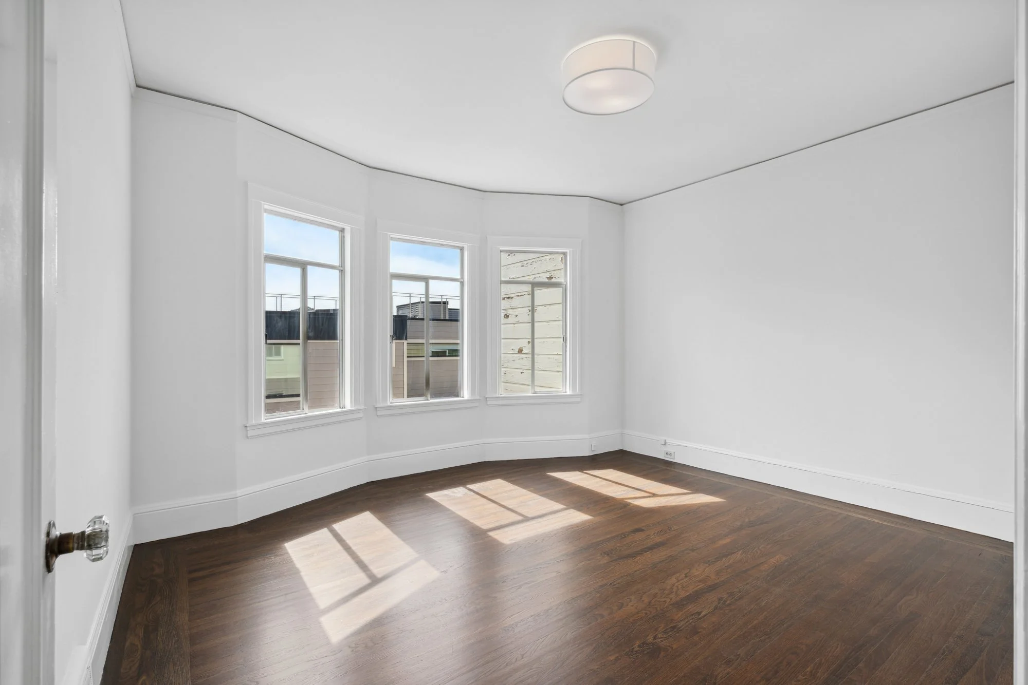 Empty room with white walls, four windows letting in natural light, dark hardwood flooring, ceiling light fixture.