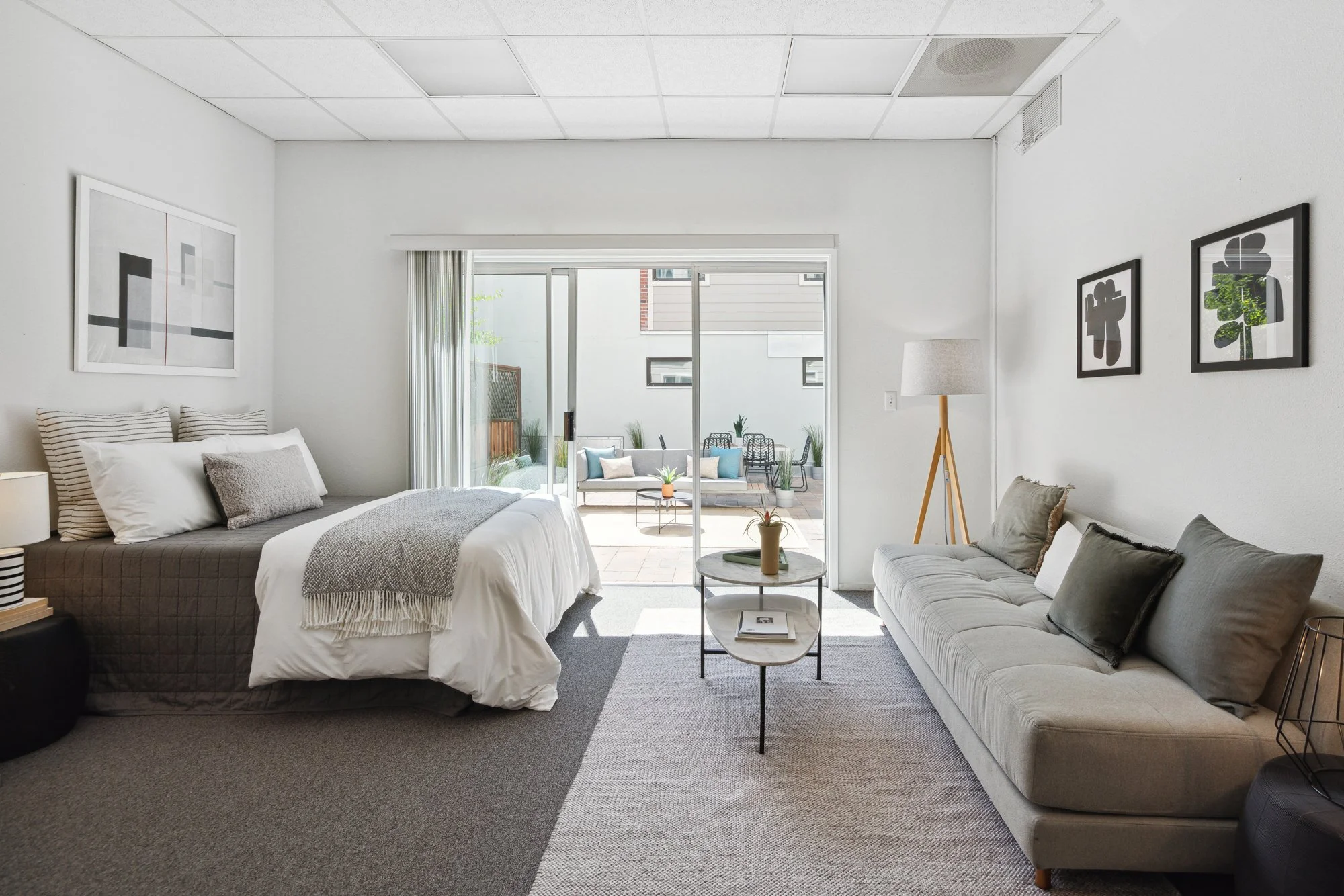 A modern living room with a large bed on the left, a beige sofa on the right, and a sliding glass door leading to an outdoor patio with chairs, a table, and potted plants. The room has white walls, framed artwork, a floor lamp, and a gray carpet.