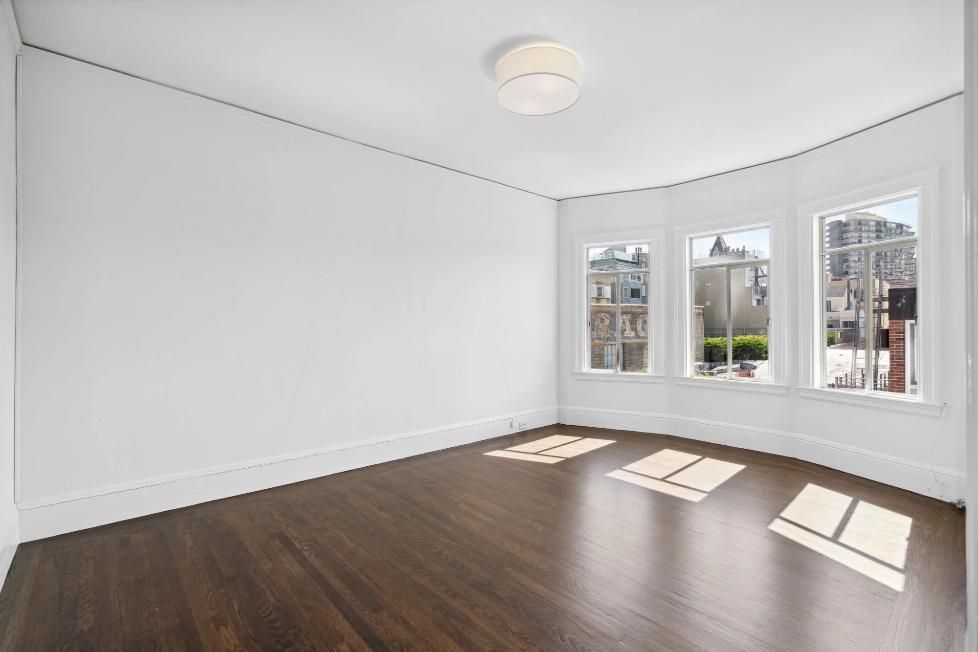 Empty room with white walls, large windows, hardwood floor, and ceiling light fixture.