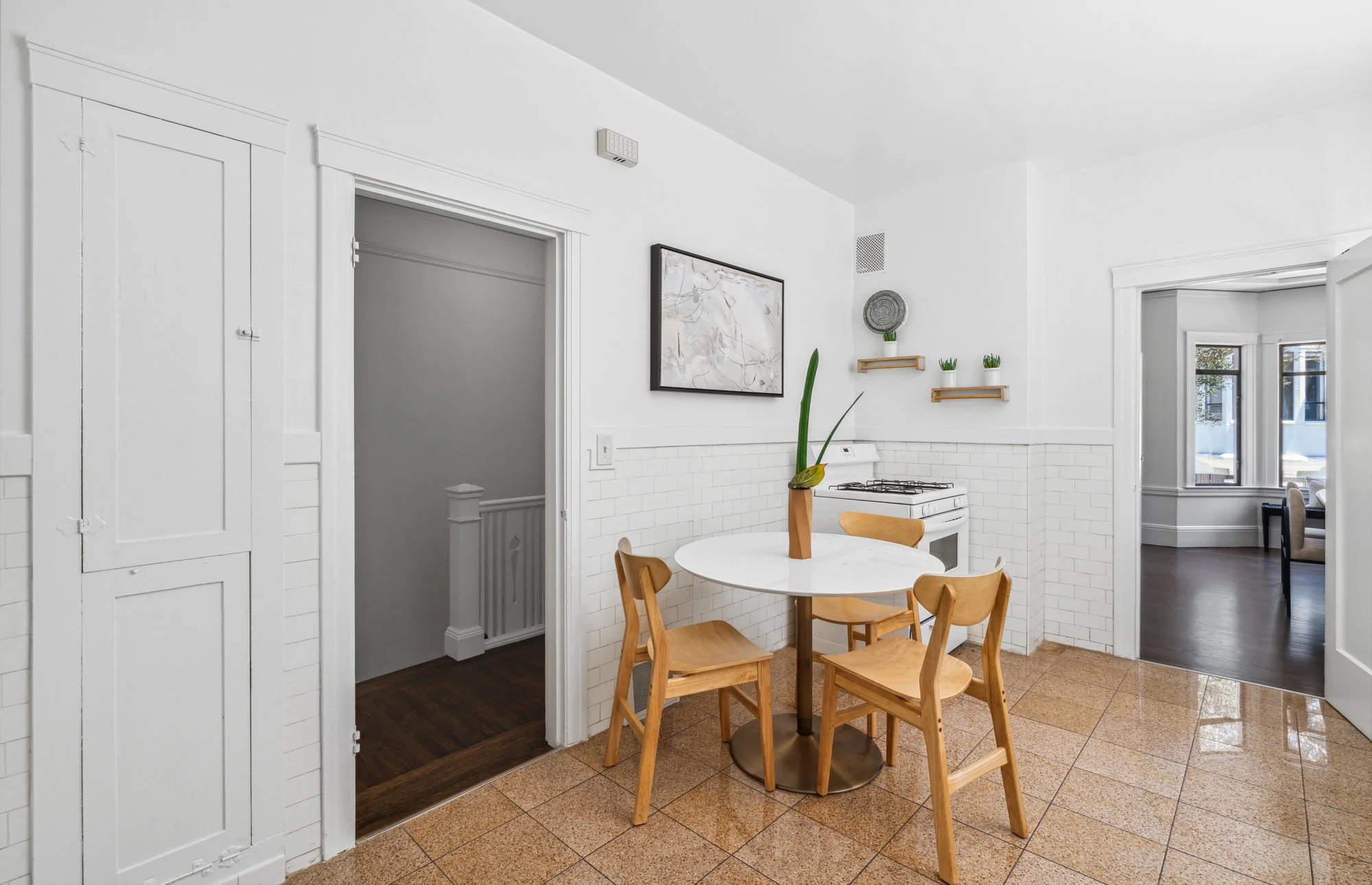 Kitchen with white walls, a dining table with four wooden chairs, a potted plant in an orange vase, wall-mounted shelves with potted plants, and a painting.