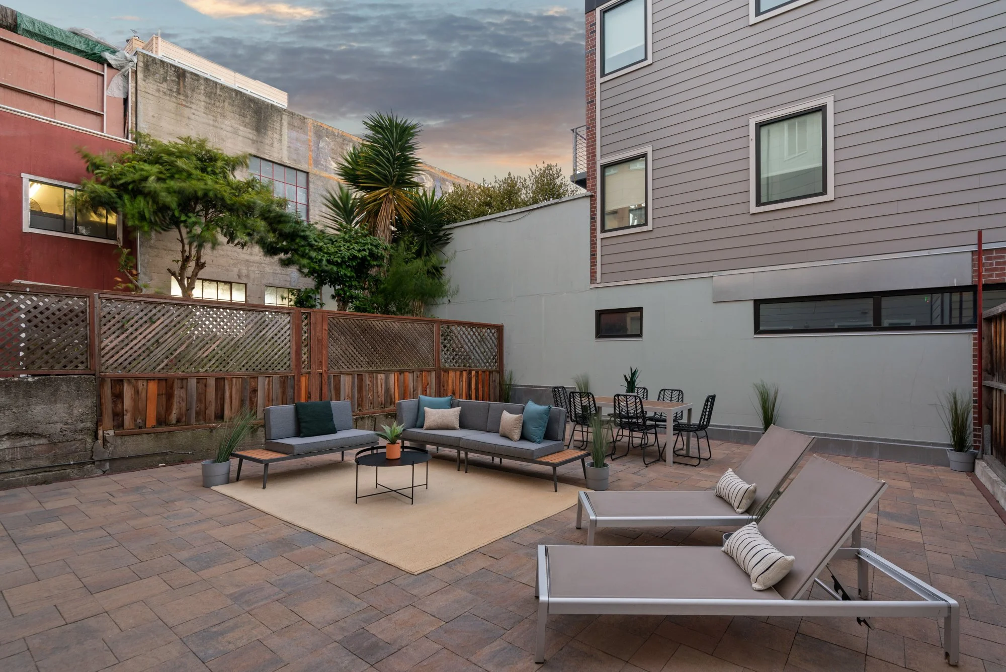 A spacious outdoor patio with modern furniture, including a gray sofa with cushions, a small round coffee table, and two reclining lounge chairs with rolled-up towels. The patio has a brick paver floor, a beige rug, and several potted plants. A woode