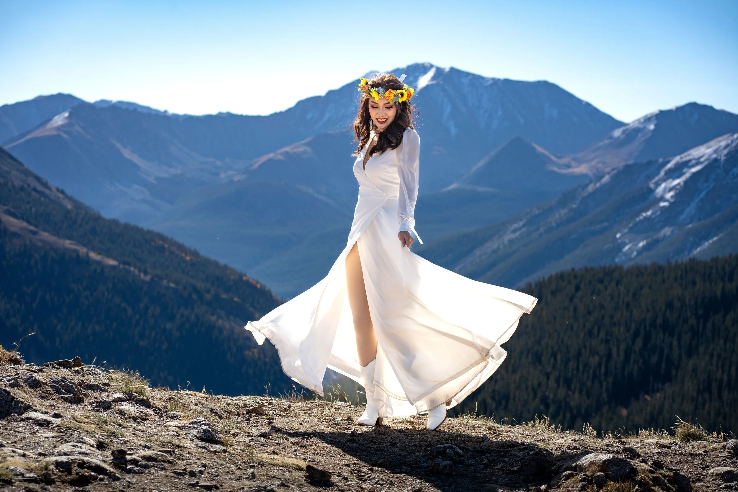 A woman in a white dress with a thigh-high slit standing on a rocky terrain outdoors, wearing a flower crown, with mountain ranges in the background, and smiling.