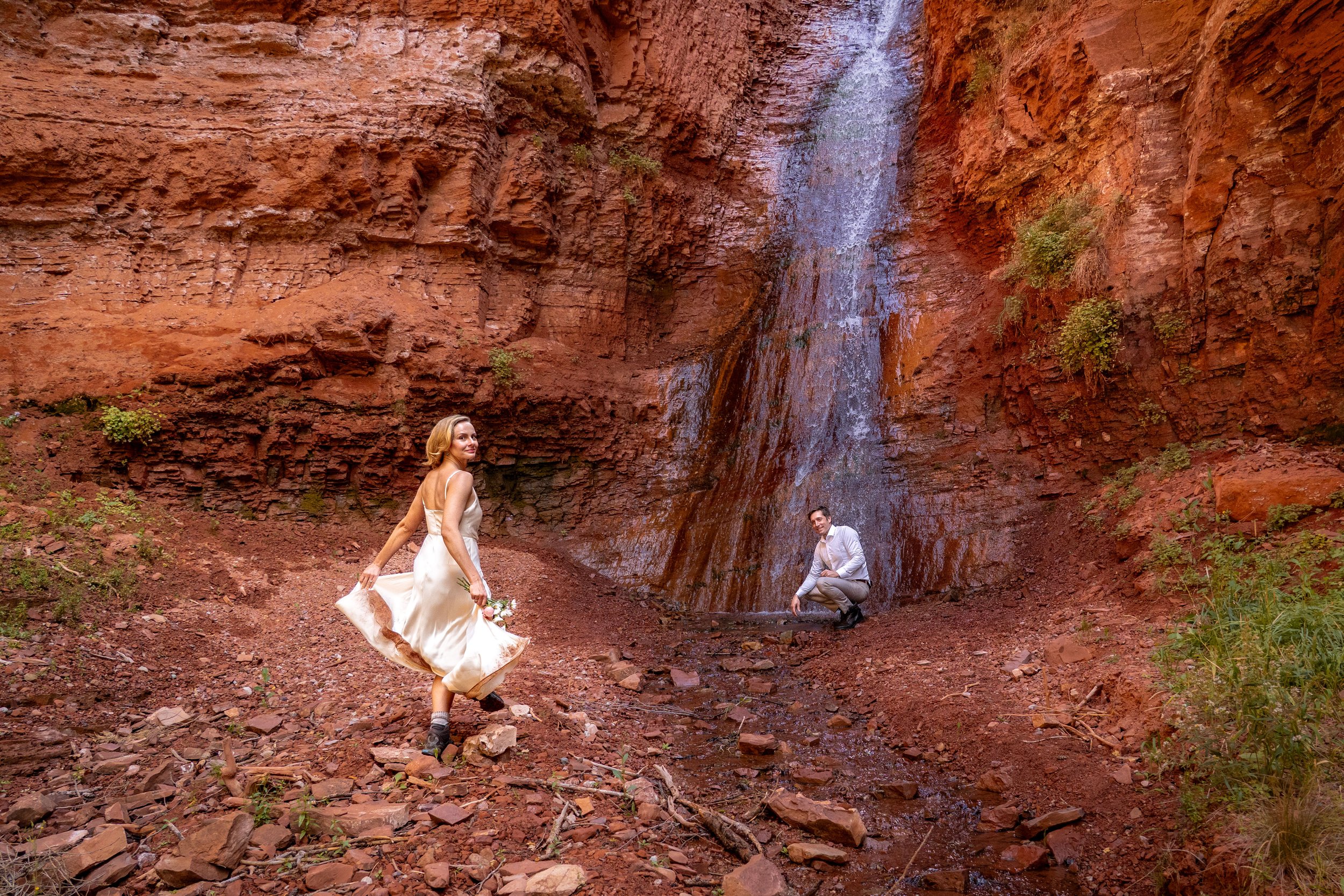 A woman in a wedding dress runs towards her groom by a waterfall.