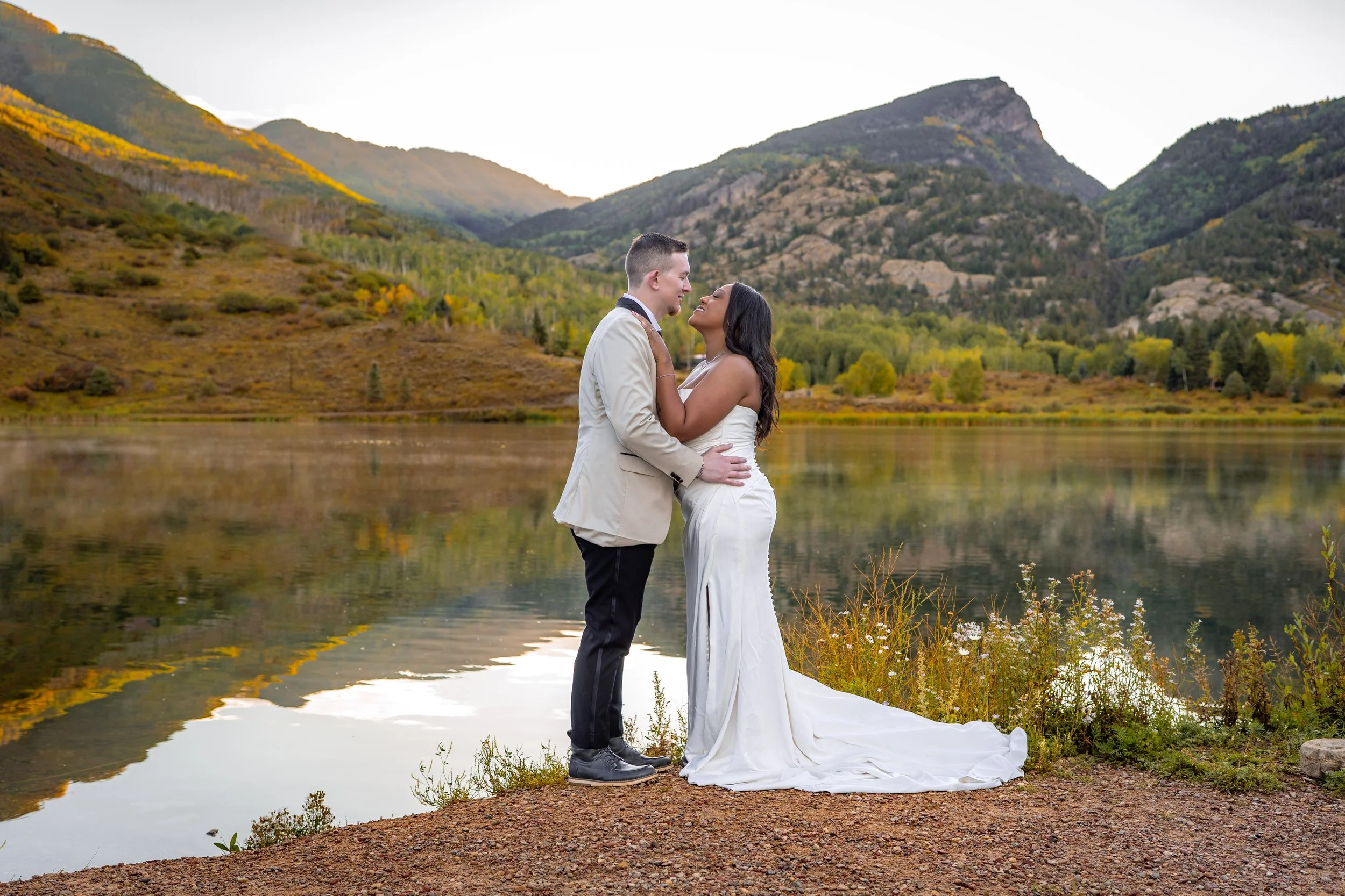 An elopement couple holds each other by a reflective alpine lake at sunrise in Colorado.