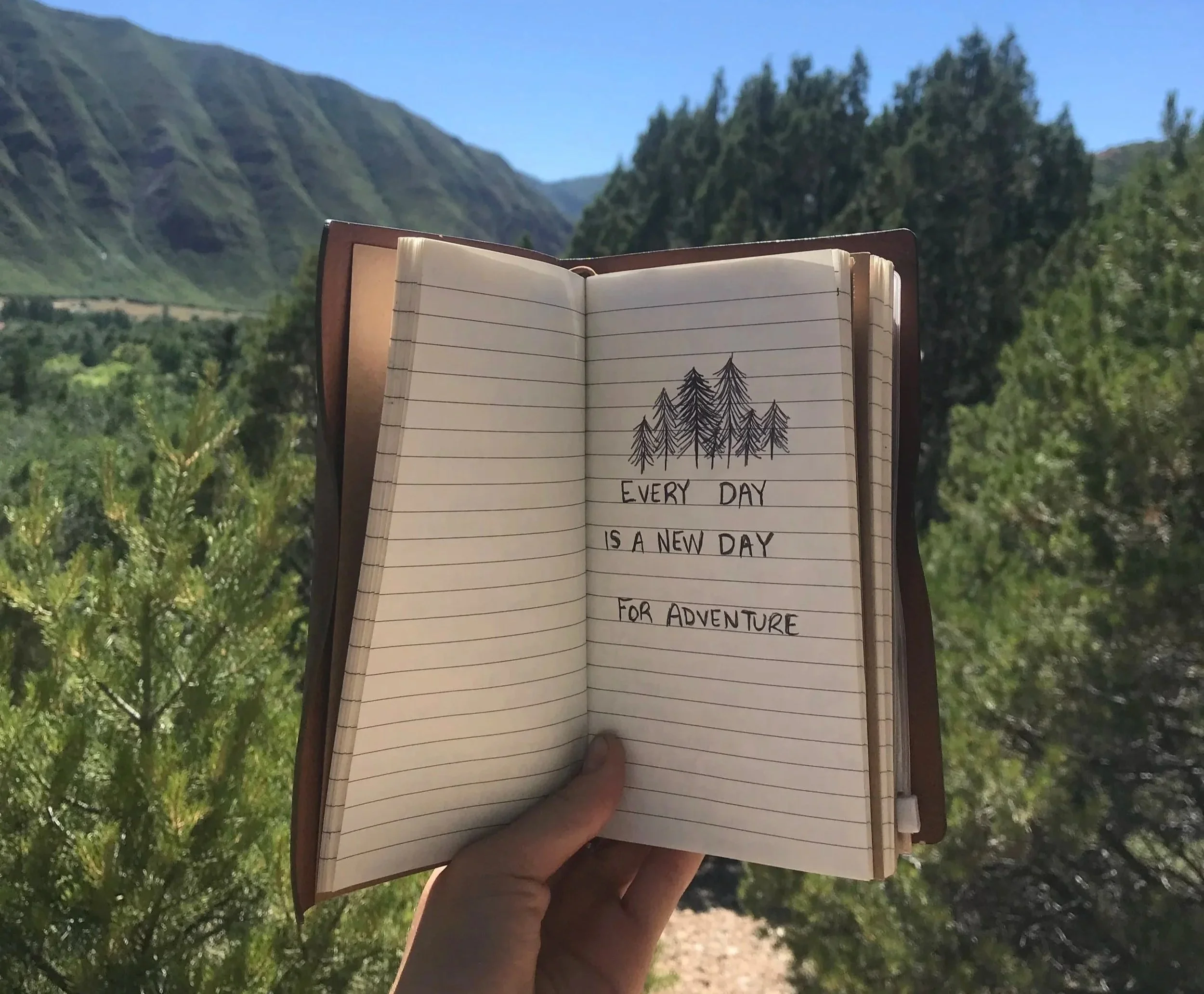 A journal held up to mountains with "Every day is a new adventure" written on it.