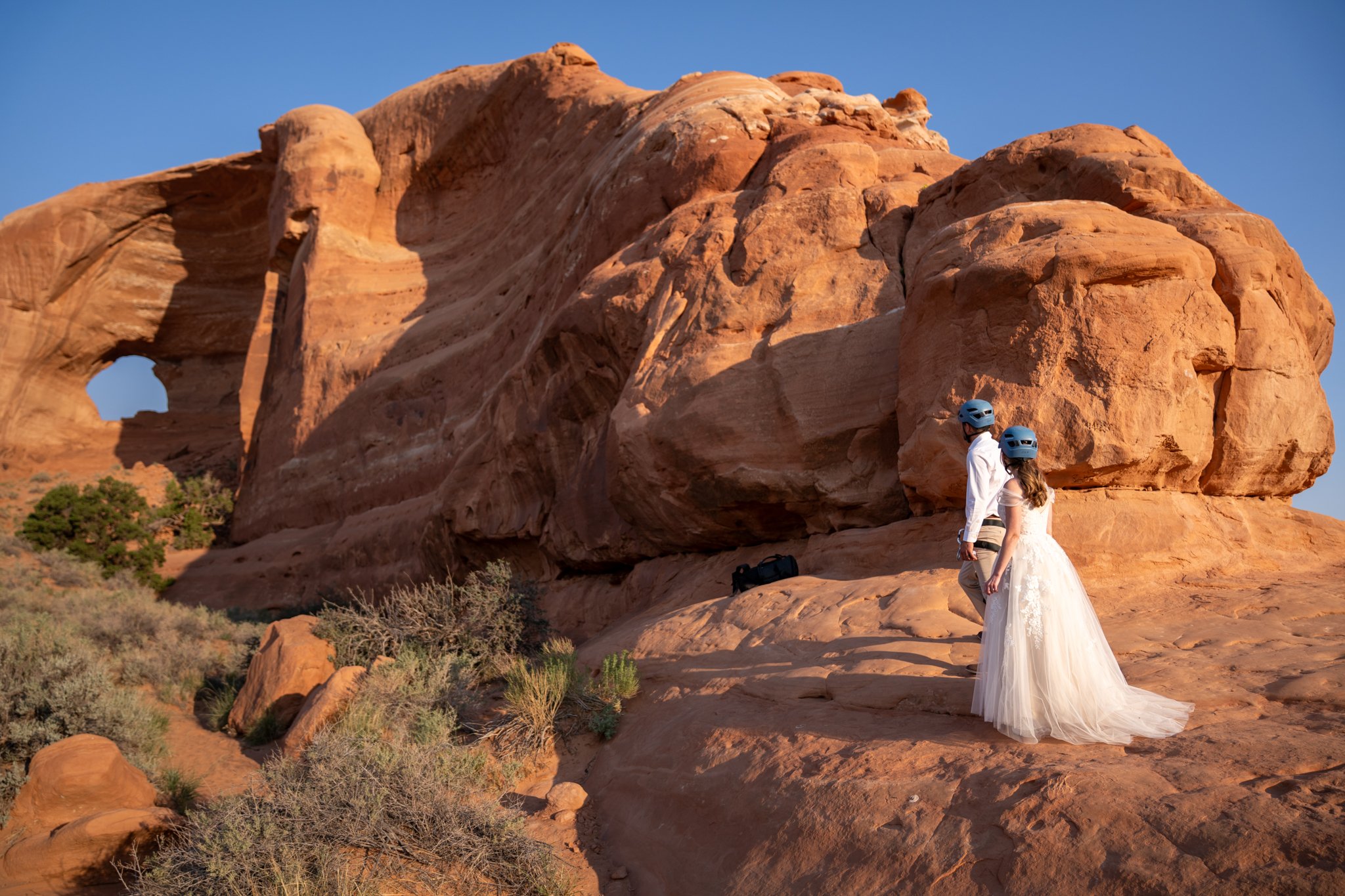 A bride and groom look at a red rock in Moab in climbing helmets.