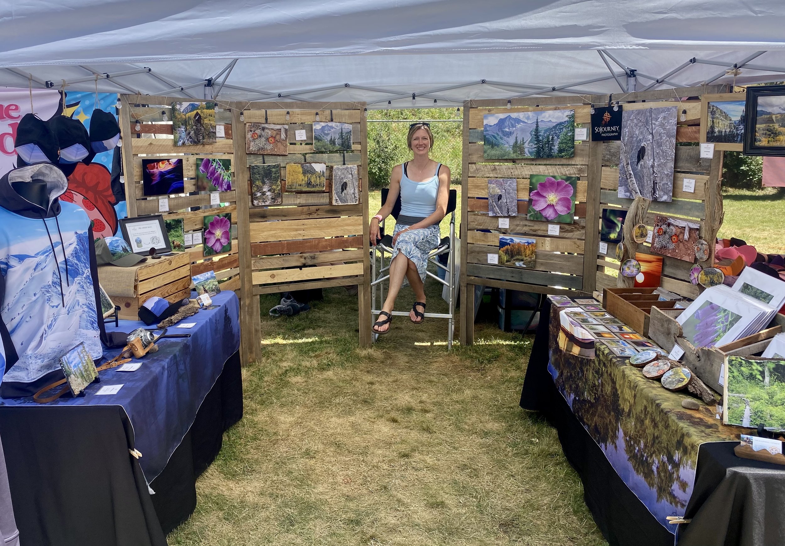Girl sitting inside tent at festival showcasing her photography work on prints, hats, and clothing.