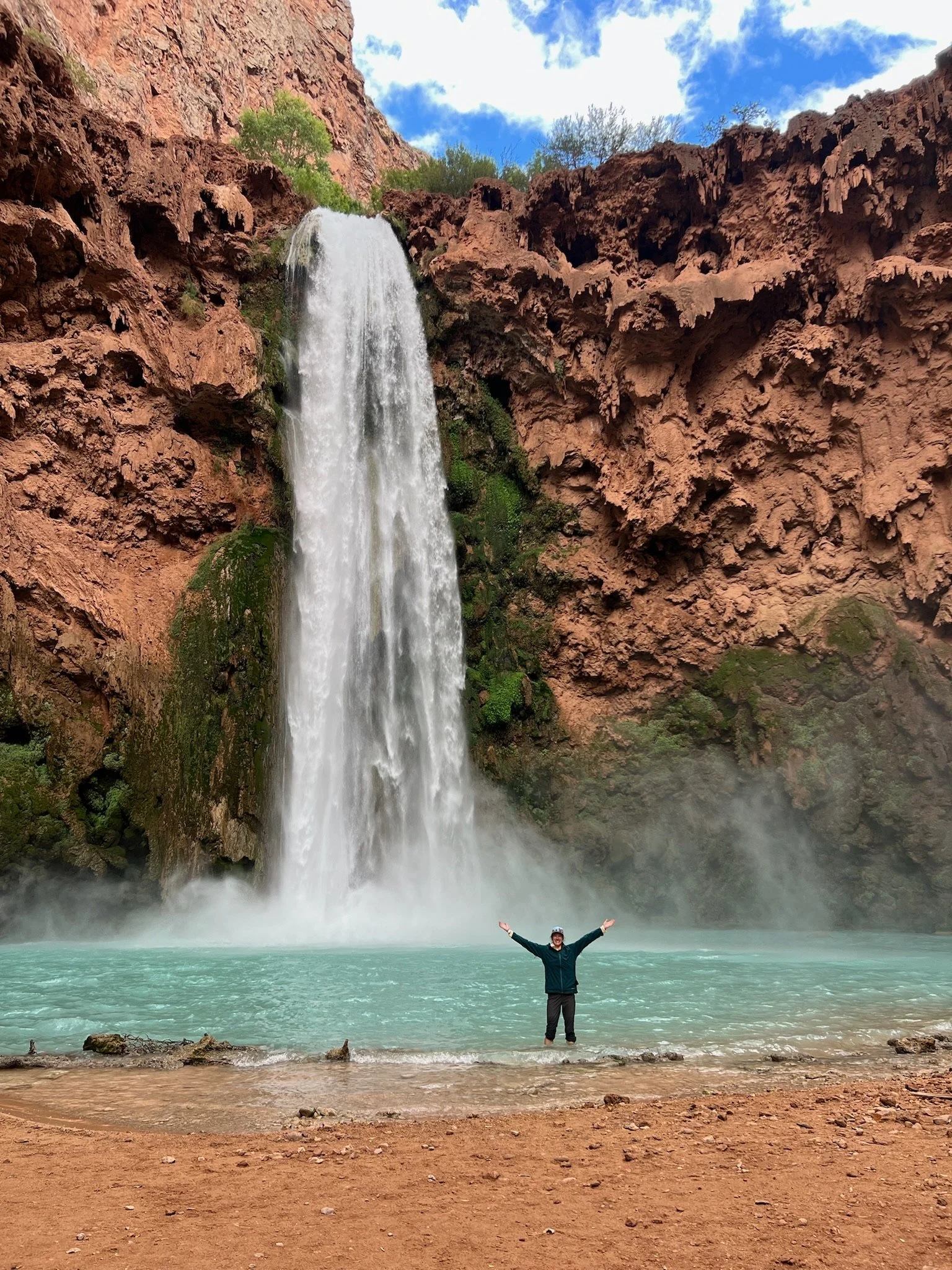 Girl joyfully holding hands in air in front of massive waterfall in Havasupai, Arizona.
