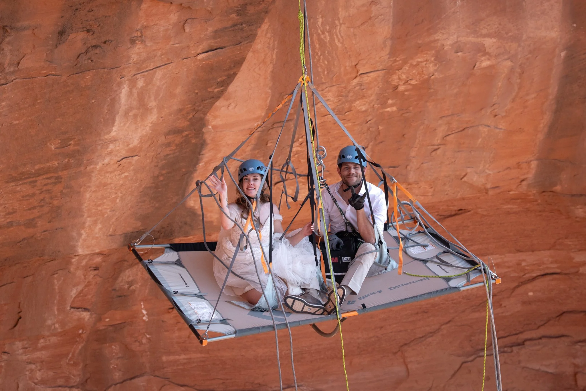 A bride and groom waive from a rock climbing suspended portaledge in Moab, Utah.