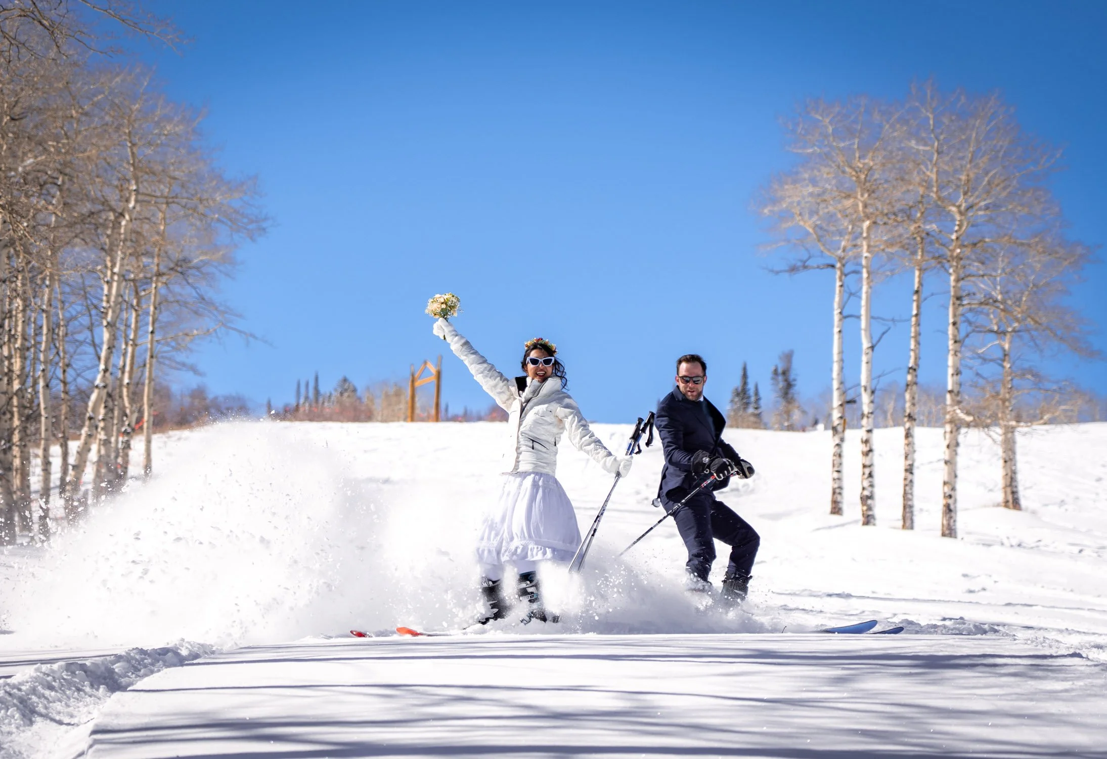 A groom skis down the mountain to spray snow onto his bride.