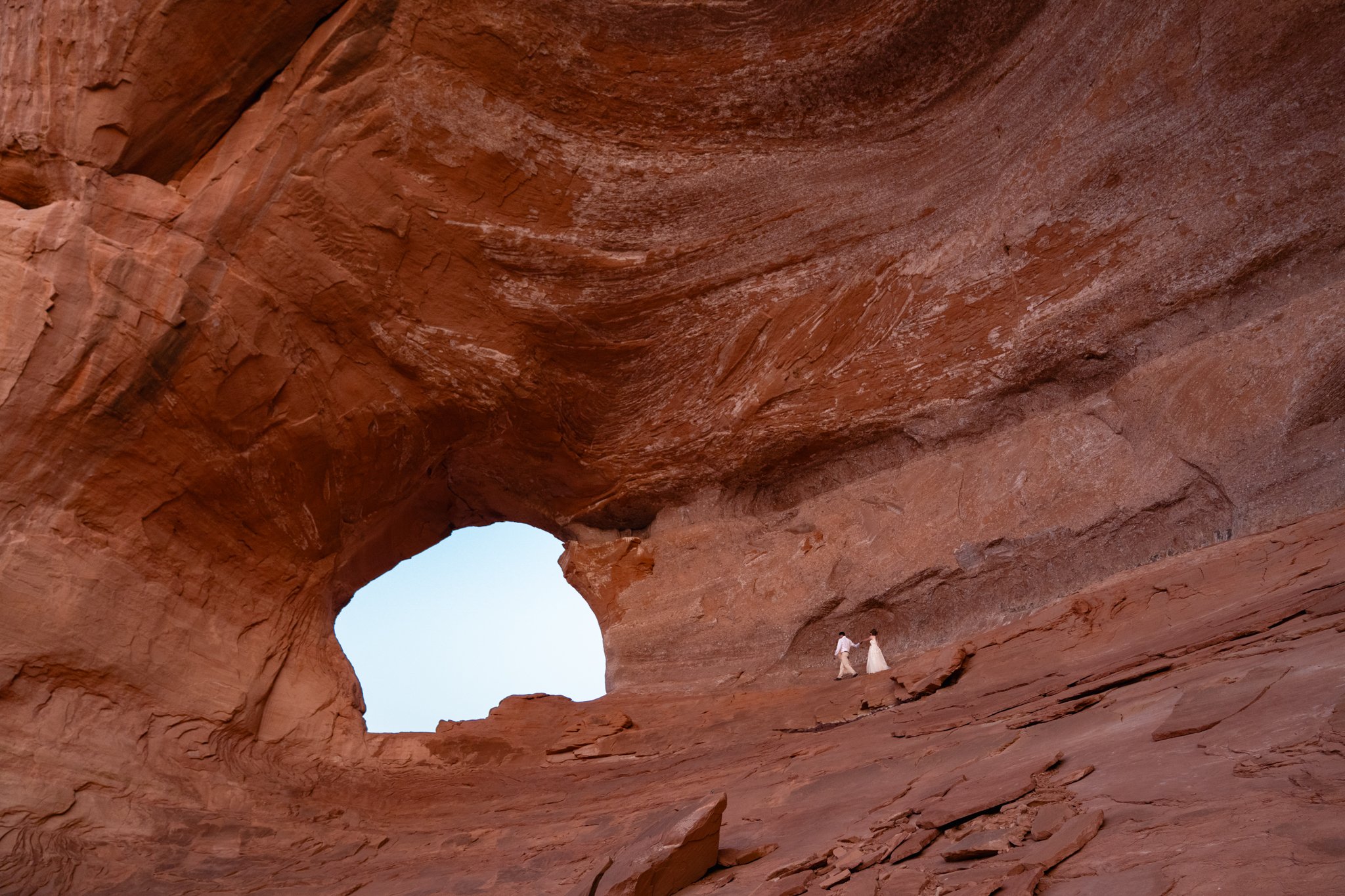 A bride and groom walk along a red rock ledge to an archway in Moab.